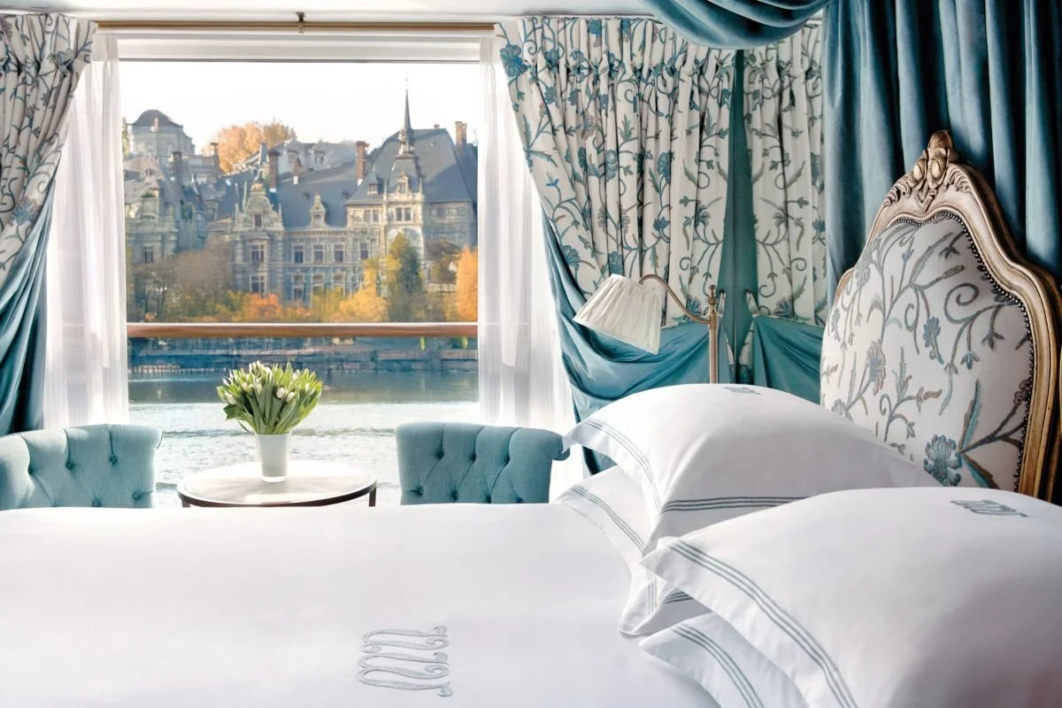 Luxurious bedroom with a bed featuring white linens and an embroidered headboard, blue and cream floral curtains, a vase of tulips on a small table, and large window overlooking a castle by a river with autumn trees.