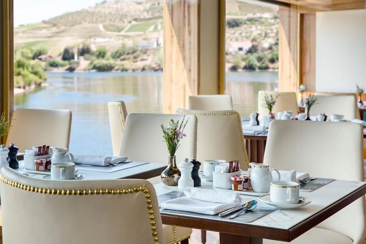 Restaurant interior with dining tables set with white cups, plates, cutlery, and small flower arrangements, overlooking a river and hills outside.