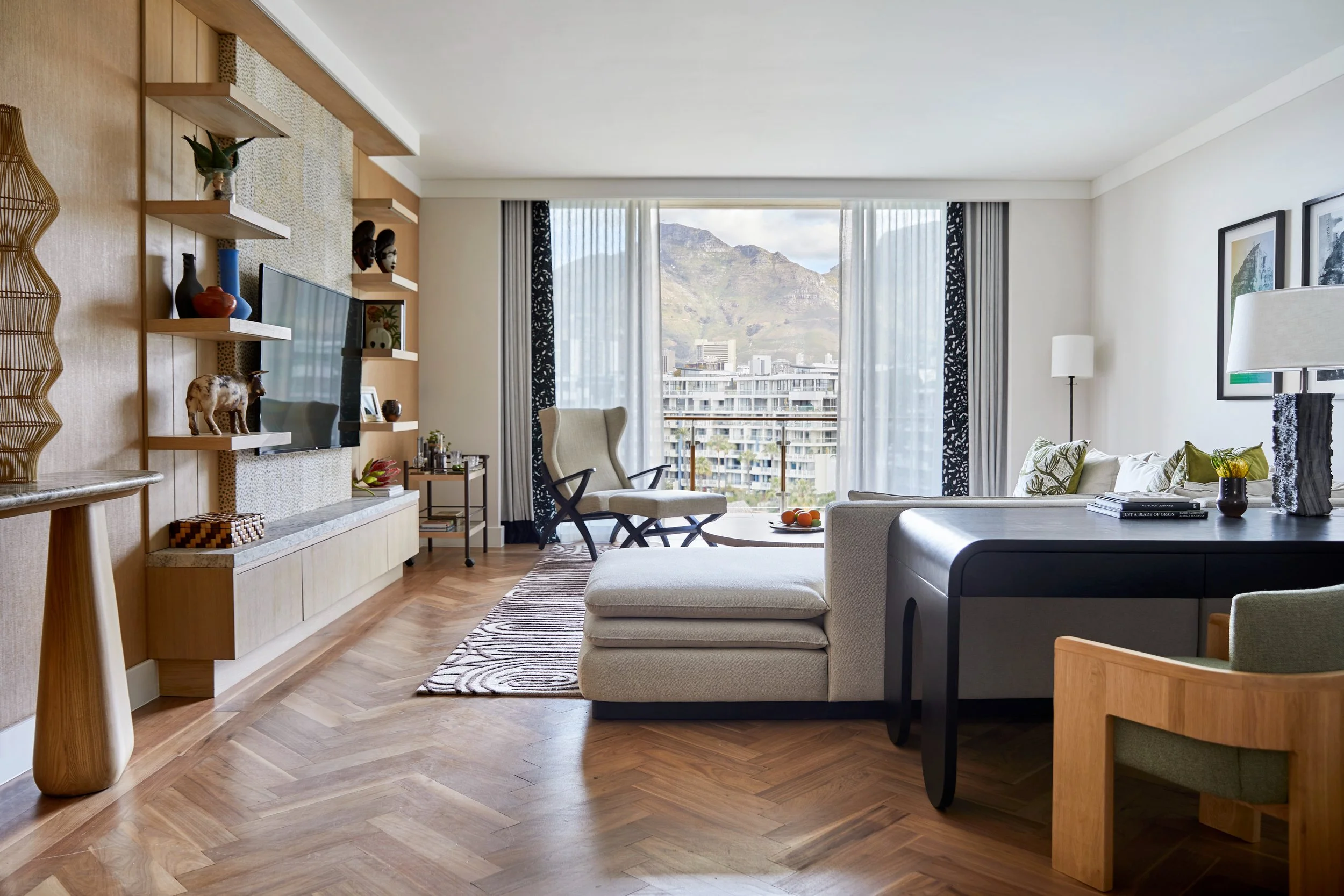 A modern living room with wooden flooring, a large window with view of mountains and cityscape, and contemporary furniture including a white sofa, beige armchair, and a black side table with decorative items and a bowl of oranges.