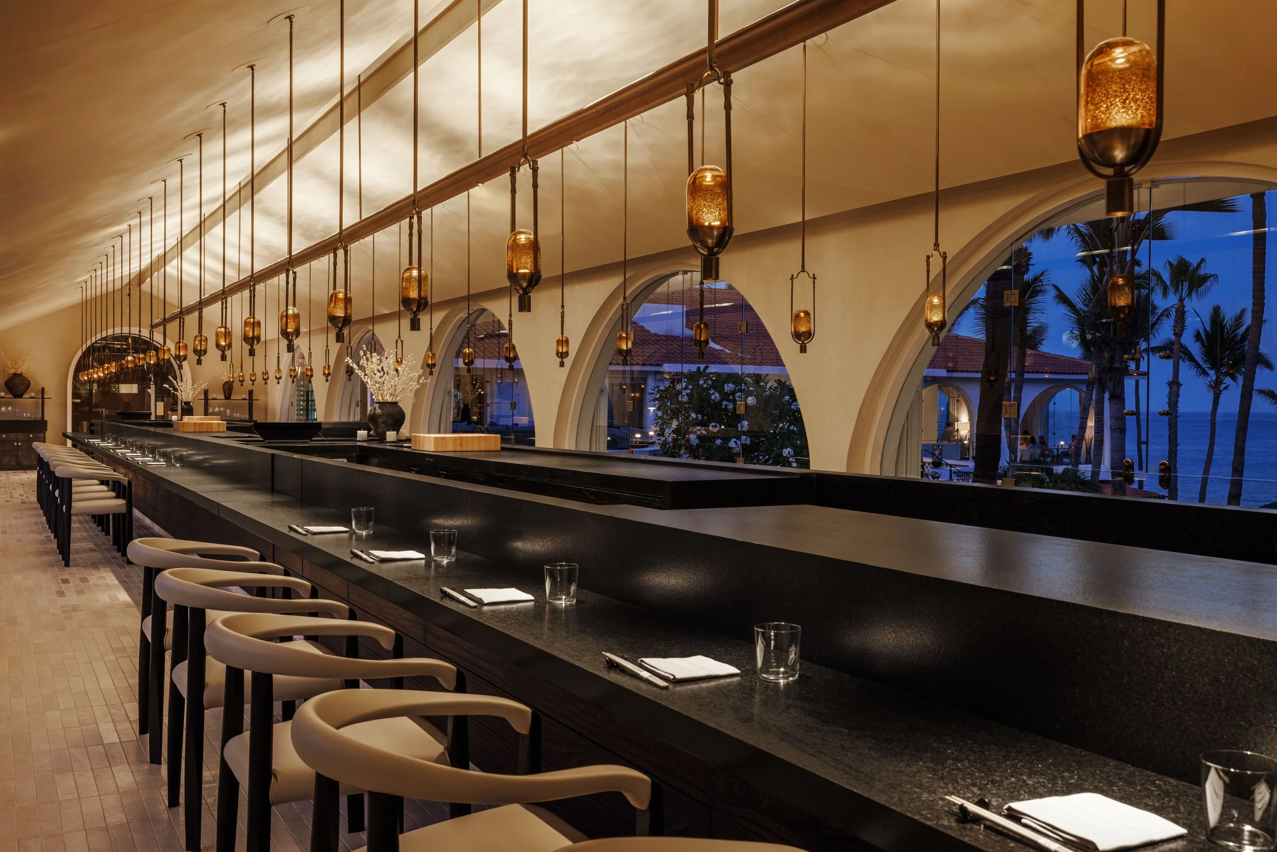 Elegant restaurant interior at dusk with a long bar counter, modern hanging pendant lights, large arched windows, and view of trees and the ocean outside.