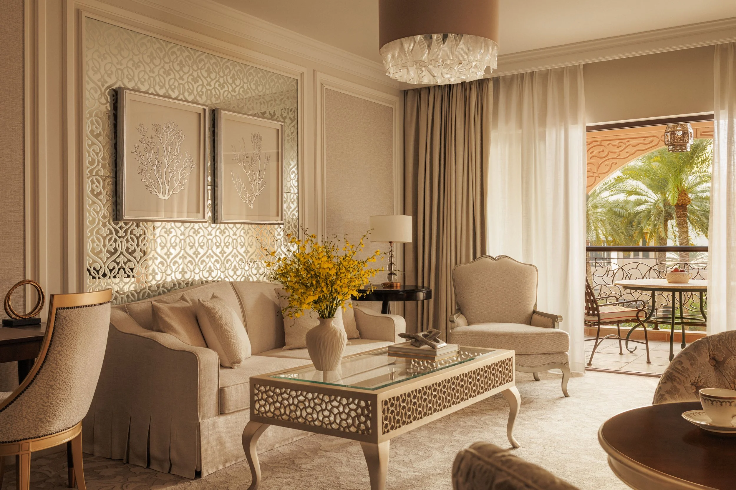 A living room with beige sofas, a glass coffee table, and decorative framed art on the wall. There are curtains and a balcony door with outdoor furniture, palm trees, and a decorative outdoor hanging lantern visible outside.