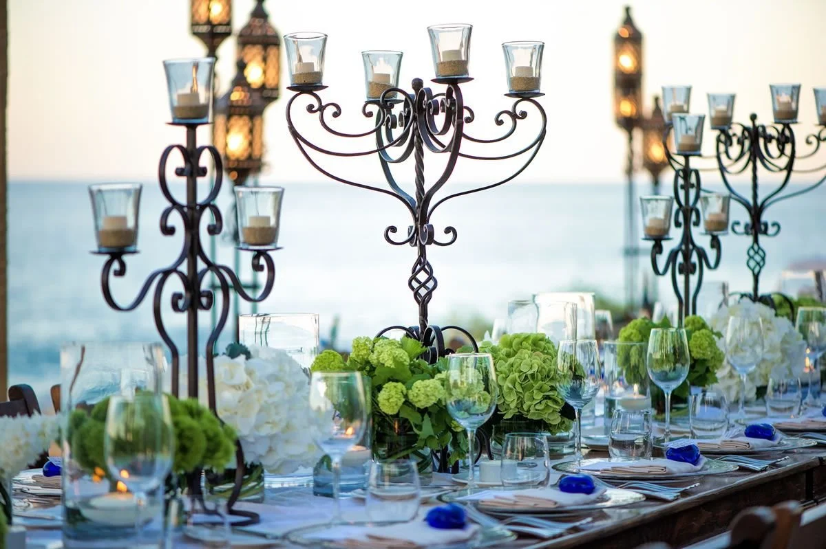 Elegant outdoor dining table decorated with white and green floral arrangements, glassware, and ornate black candle holders, with a view of the ocean in the background.