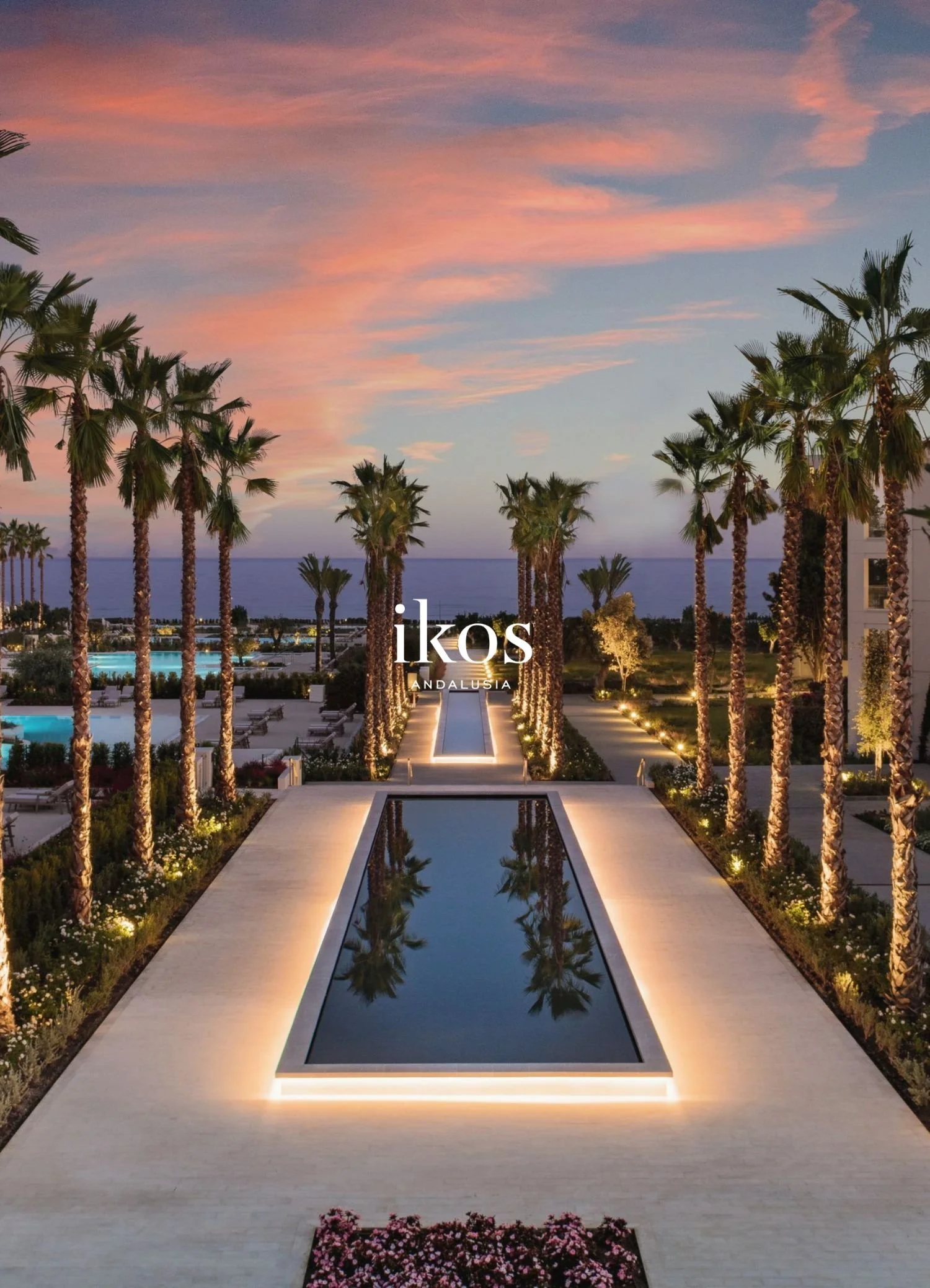 A luxurious resort with a symmetrical infinity pool reflecting palm trees, illuminated pathway, and a colorful sunset sky above the ocean in the background.