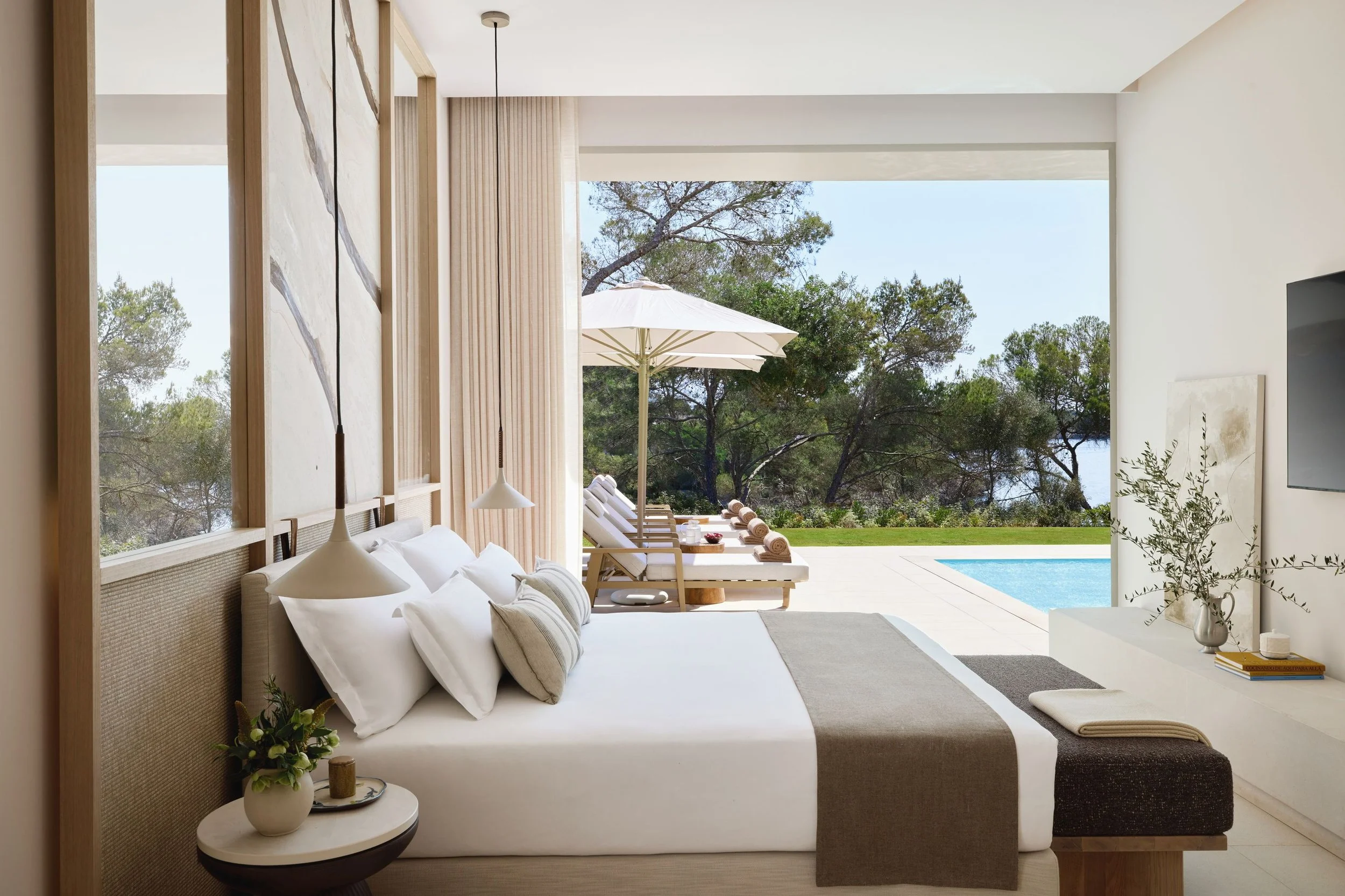 A modern bedroom with a large bed, white bedding, and multiple pillows. The room has large open windows showing trees and an outdoor patio with lounge chairs and a white umbrella. There is a small side table with a potted plant and decorative items, and a flat-screen TV on the wall.