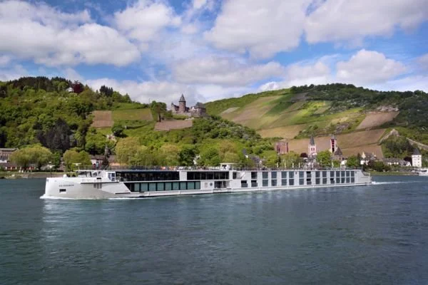 Large white modern cruise ship sailing on a river with green hills and buildings in the background.