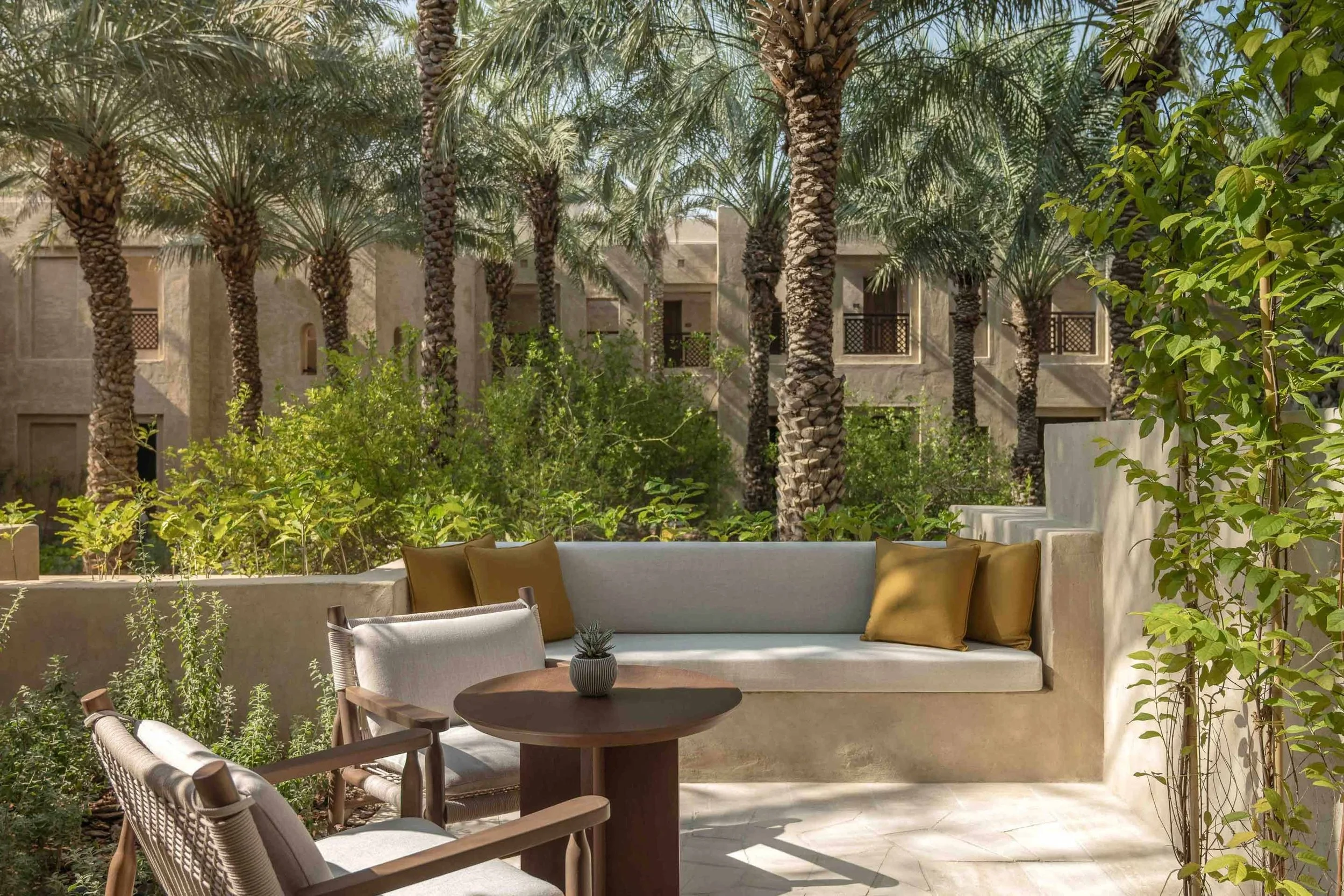 A patio with outdoor furniture including a cushioned sofa with gold pillows, a round wooden table, and chairs, surrounded by green plants and trees in front of a building with palm trees and windows.