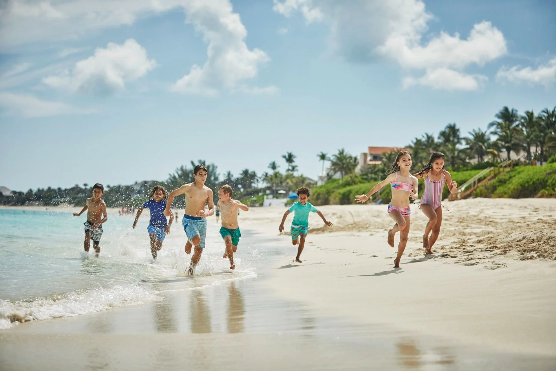 Children running and playing along the shoreline at the beach on a sunny day, with palm trees and residential houses in the background.