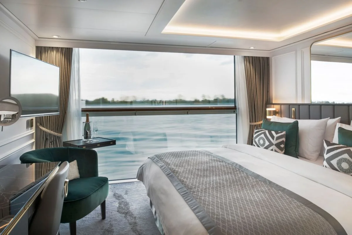 A luxurious hotel room with a large bed, decorative pillows, a window view of water, a television, a small table with a champagne bottle and glasses, and elegant curtains.