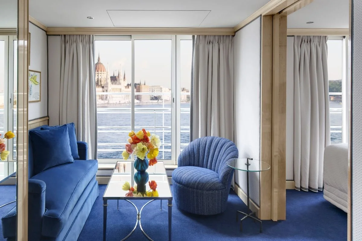 Interior view of a luxurious room with a blue velvet sofa, a matching blue armchair, a glass top table with colorful flowers, large windows overlooking a river and historic buildings.