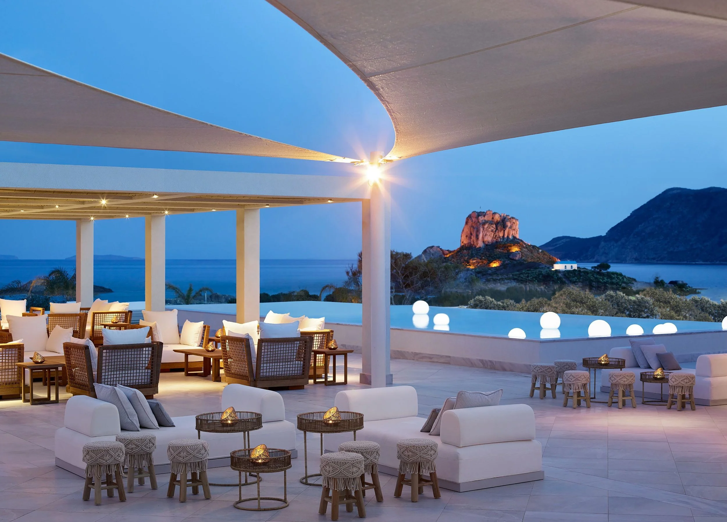 Elegant outdoor lounge area with white cushioned seating, wooden chairs, small round tables with decorative lanterns, and a covered area with white fabric sails against a backdrop of water, mountains, and a small white building at sunset.