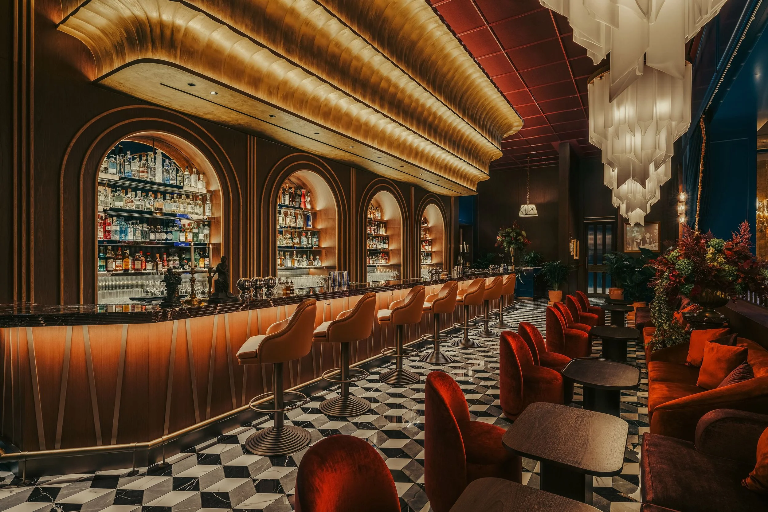 A stylish bar with a marble countertop, surrounded by red velvet chairs and high cream-colored bar stools. The bar has arched wooden shelving filled with various bottles of alcohol. The ceiling features ornate lighting fixtures and decorative woven panels, with a checkered black-and-white floor and a floral arrangement in the corner.