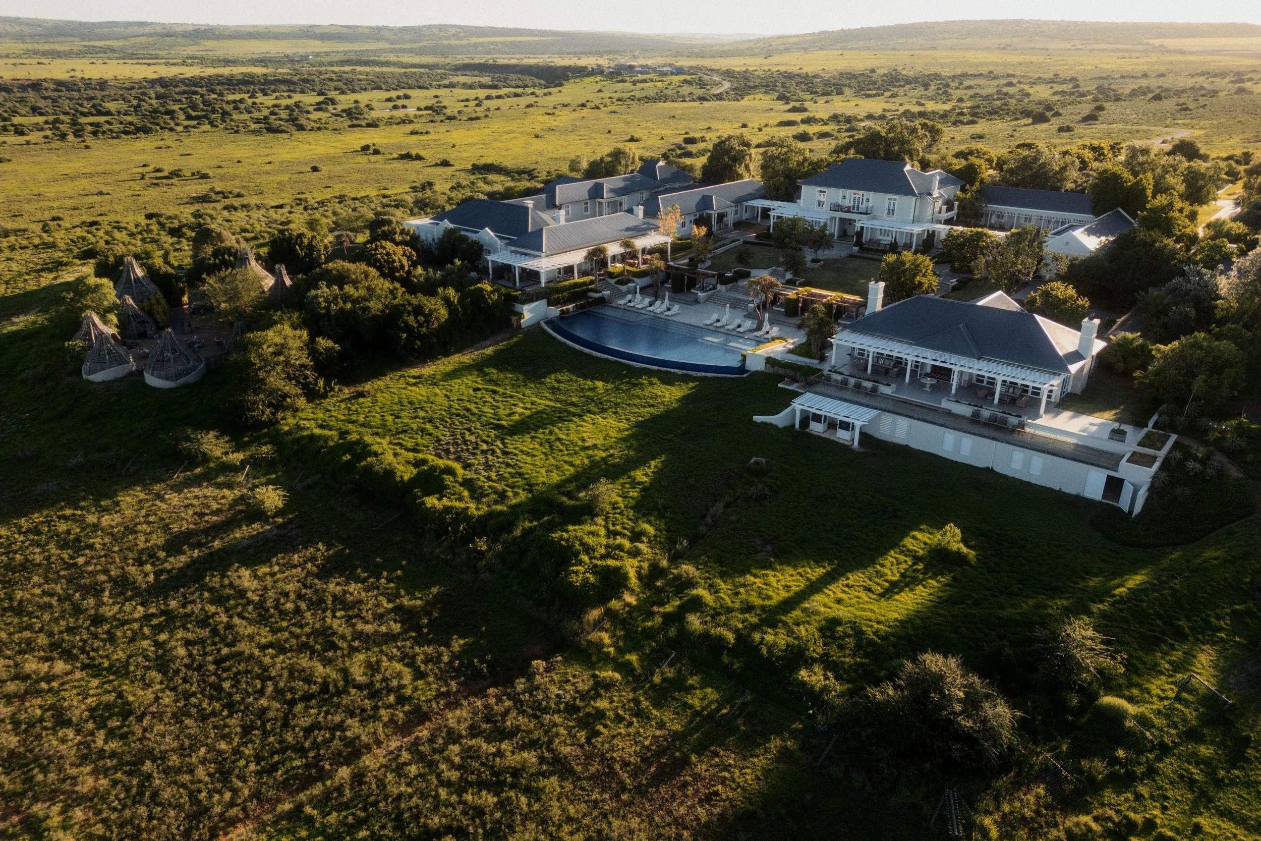 A large white house with a swimming pool and surrounding trees in a rural area with open fields in the background.