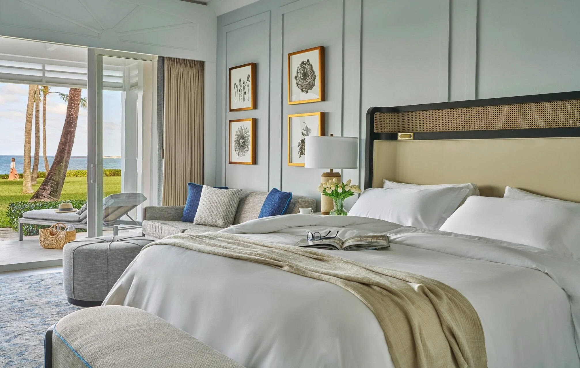 Hotel room with a large bed, nightstand, lamp, framed botanical art on the wall, and a view of palm trees and the ocean through a glass door.