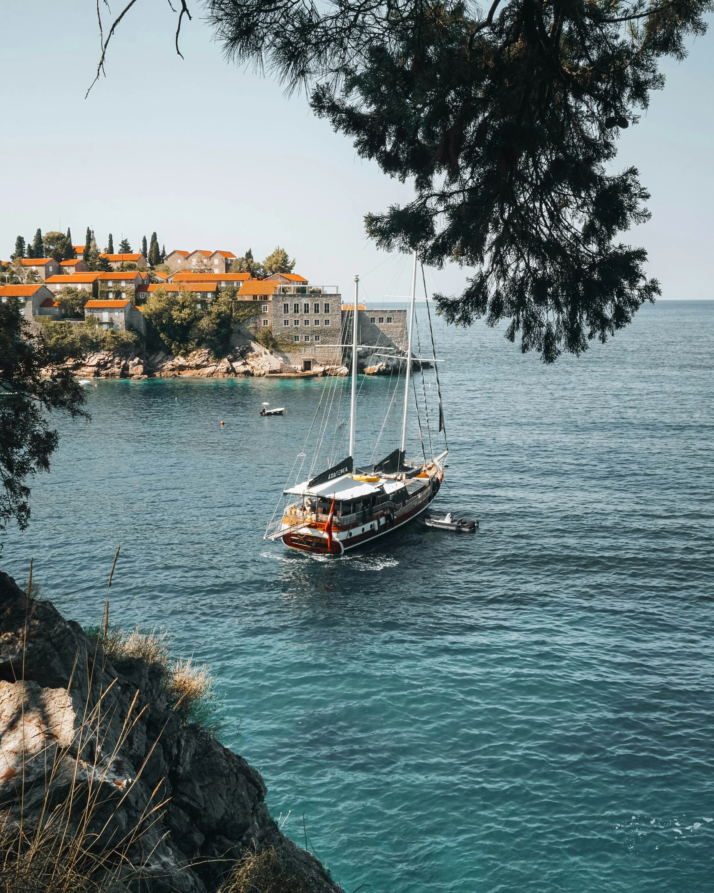 A scenic coastal scene with a sailboat in the water, rocky shoreline, trees, and houses with red-tiled roofs along the hilltop, and a large pine tree branch hanging overhead.