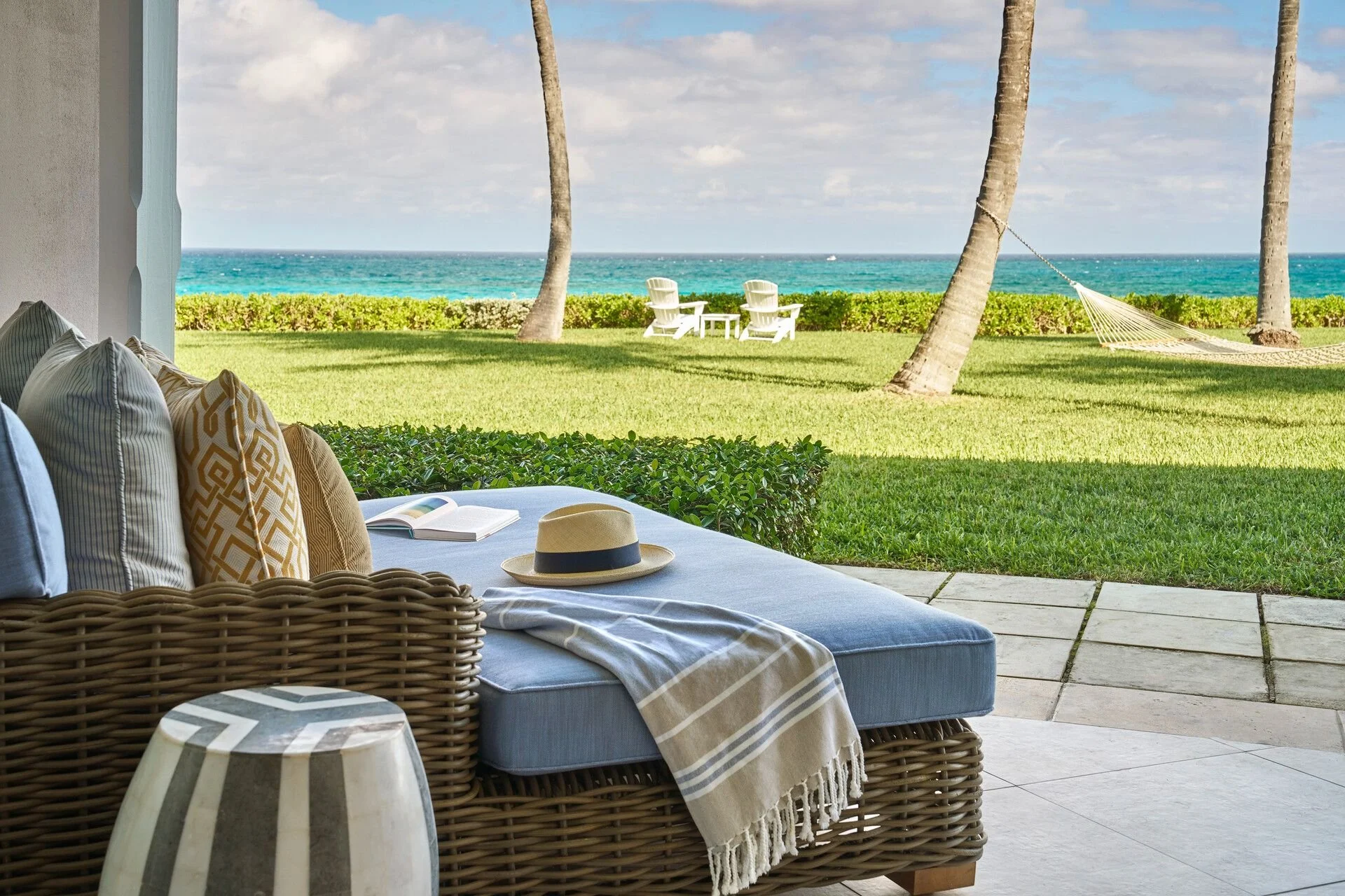 A patio view overlooking a green lawn with two palm trees, a hammock, two white Adirondack chairs, and an ocean in the background under a partly cloudy sky. Inside, there is a wicker sofa with cushions, a hat, and an open book on a table.