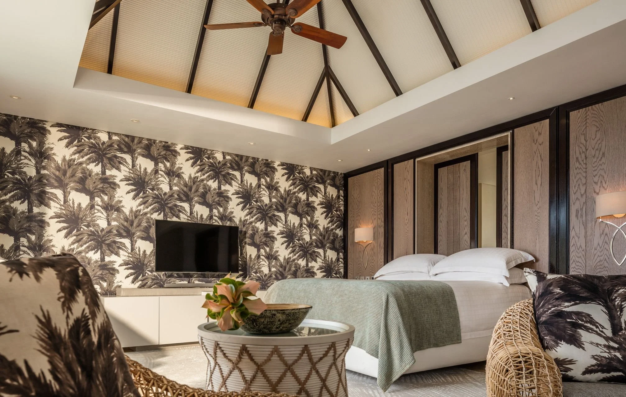 A bedroom with a high vaulted ceiling, ceiling fan, palm tree wallpaper, wooden wall panels, and a bed with white bedding. There is a flat-screen TV and a decorative side table with a plant.