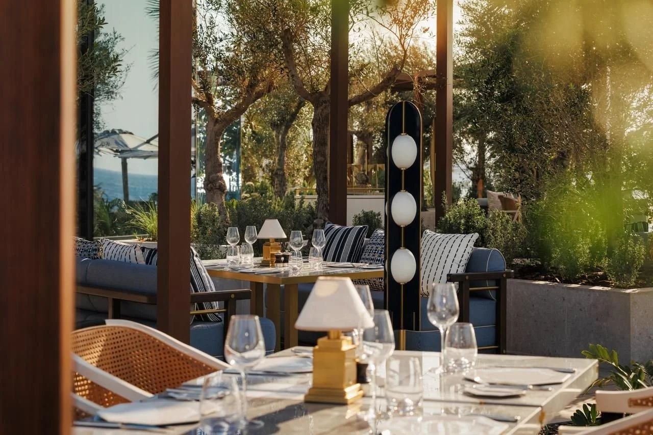 Elegant outdoor restaurant patio with tables set with wine glasses, plates, and lamps, surrounded by trees and greenery.