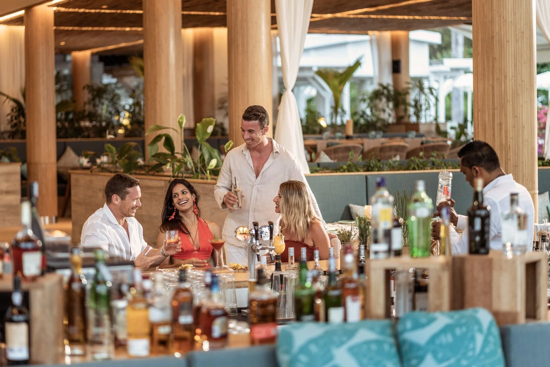 A group of people socializing at a bar or restaurant with bartenders serving drinks, set in a modern, tropical-inspired decor featuring wooden beams, greenery, and large windows.