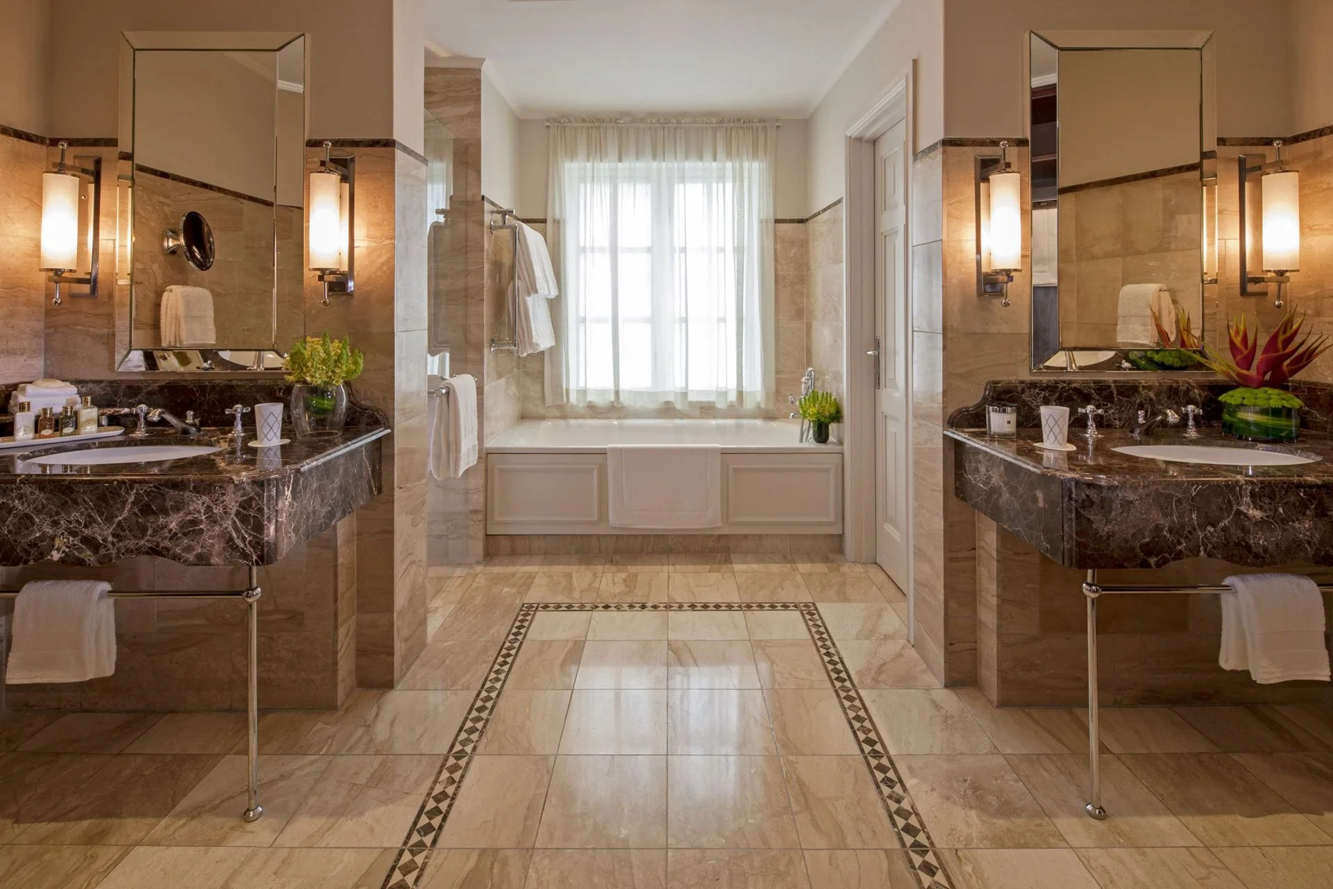 A luxurious bathroom with two marble vanities, each with a mirror and wall-mounted light fixture. In the center, there's a bathtub with a window covered by sheer curtains behind it, and two plants on either side of the bathtub. The floor and walls are tiled in beige with decorative border patterns.