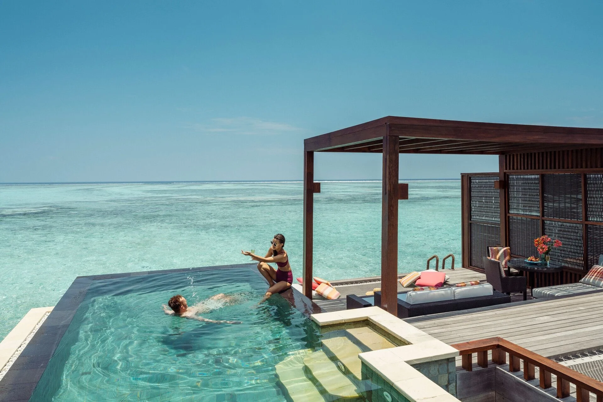 A woman sitting on the edge of a swimming pool, with a person in the water, overlooking the ocean, with a modern cabana with outdoor furniture nearby.