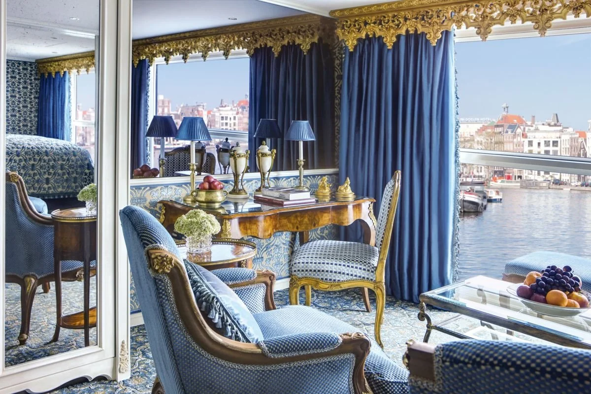 Luxurious hotel room with blue and gold decor, a large window overlooking a canal with boats, and elegant furniture including a desk with blue lamps, a sofa, chairs, and a glass coffee table with fruit.