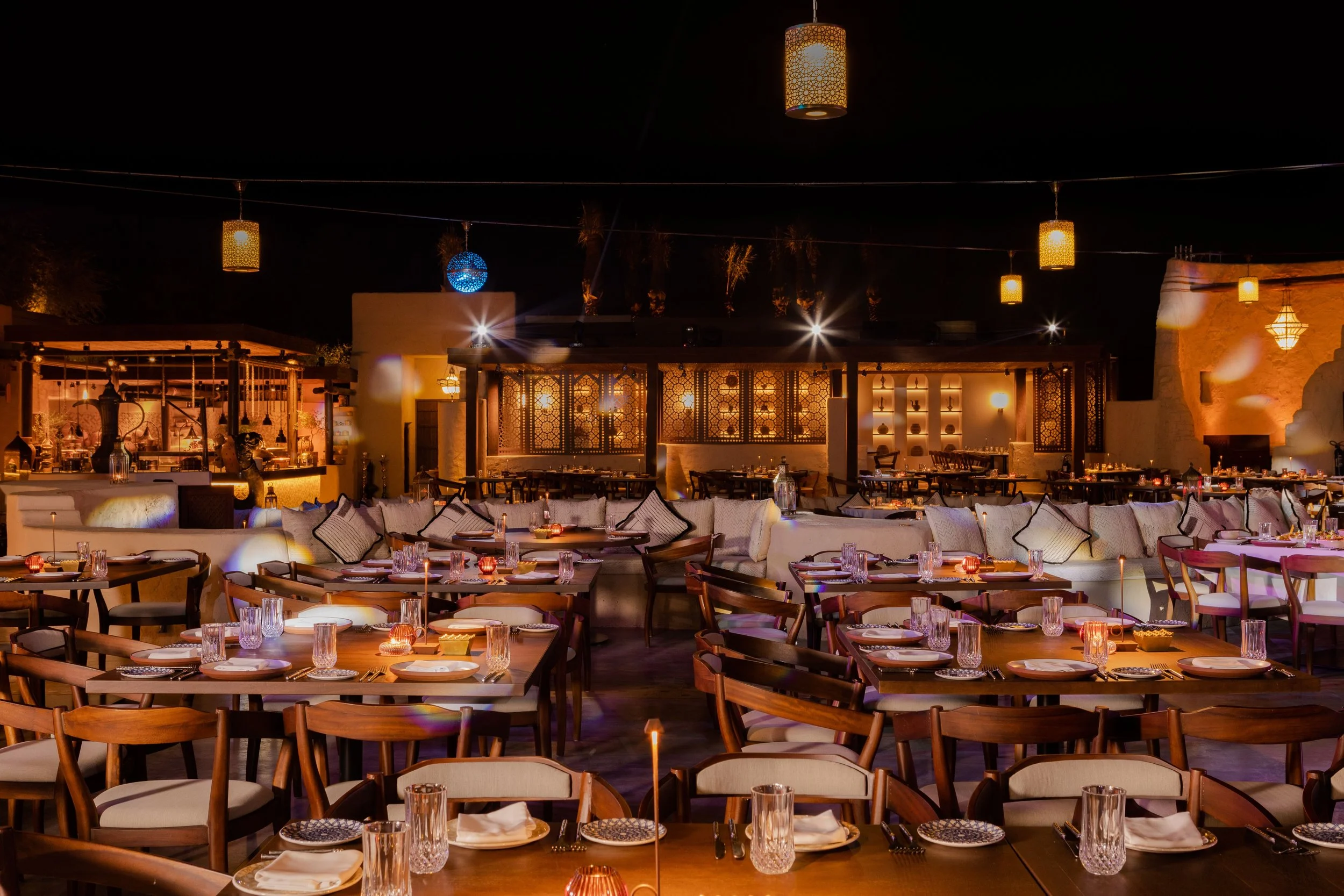 A nighttime outdoor dining area with wooden tables and chairs, decorated with candles, surrounded by beige sofas with cushions, and illuminated by hanging lanterns and warm lighting.