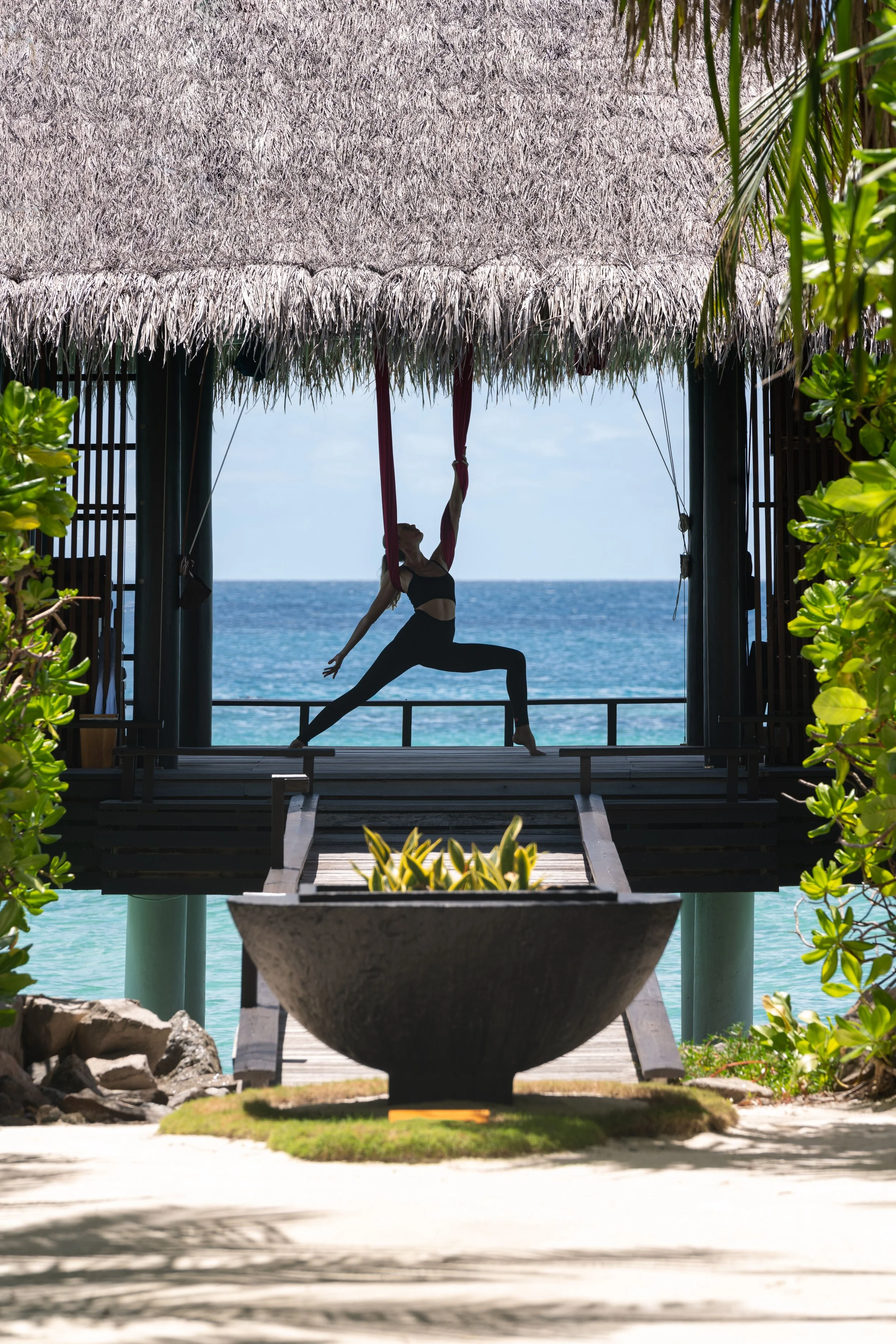 A woman practicing aerial yoga on pink fabric hanging from a wooden structure with thatched roof, overlooking the ocean on a sunny day, framed by greenery.