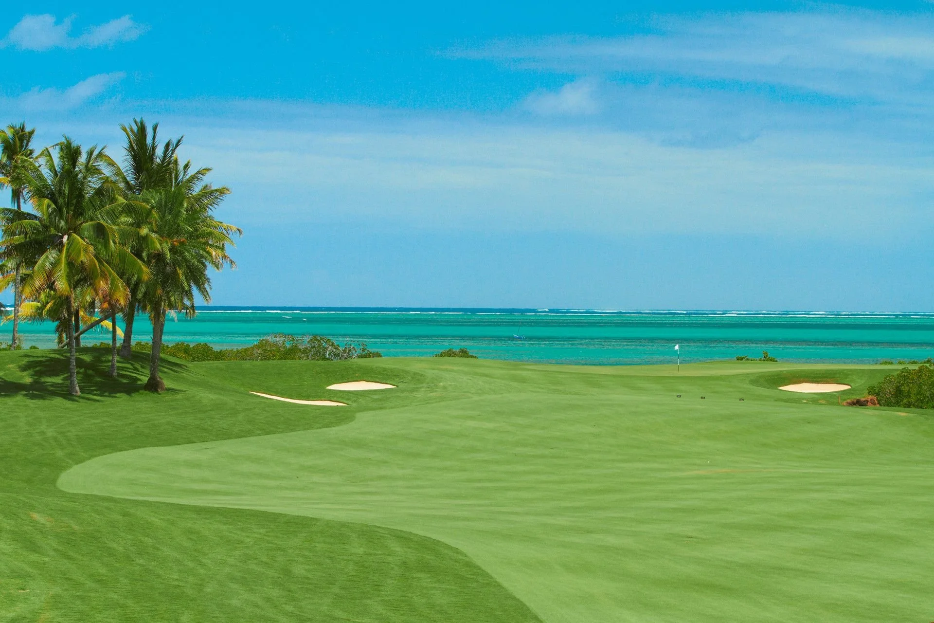 A golf course with sand traps, lush green grass, and palm trees overlooking the ocean on a sunny day.