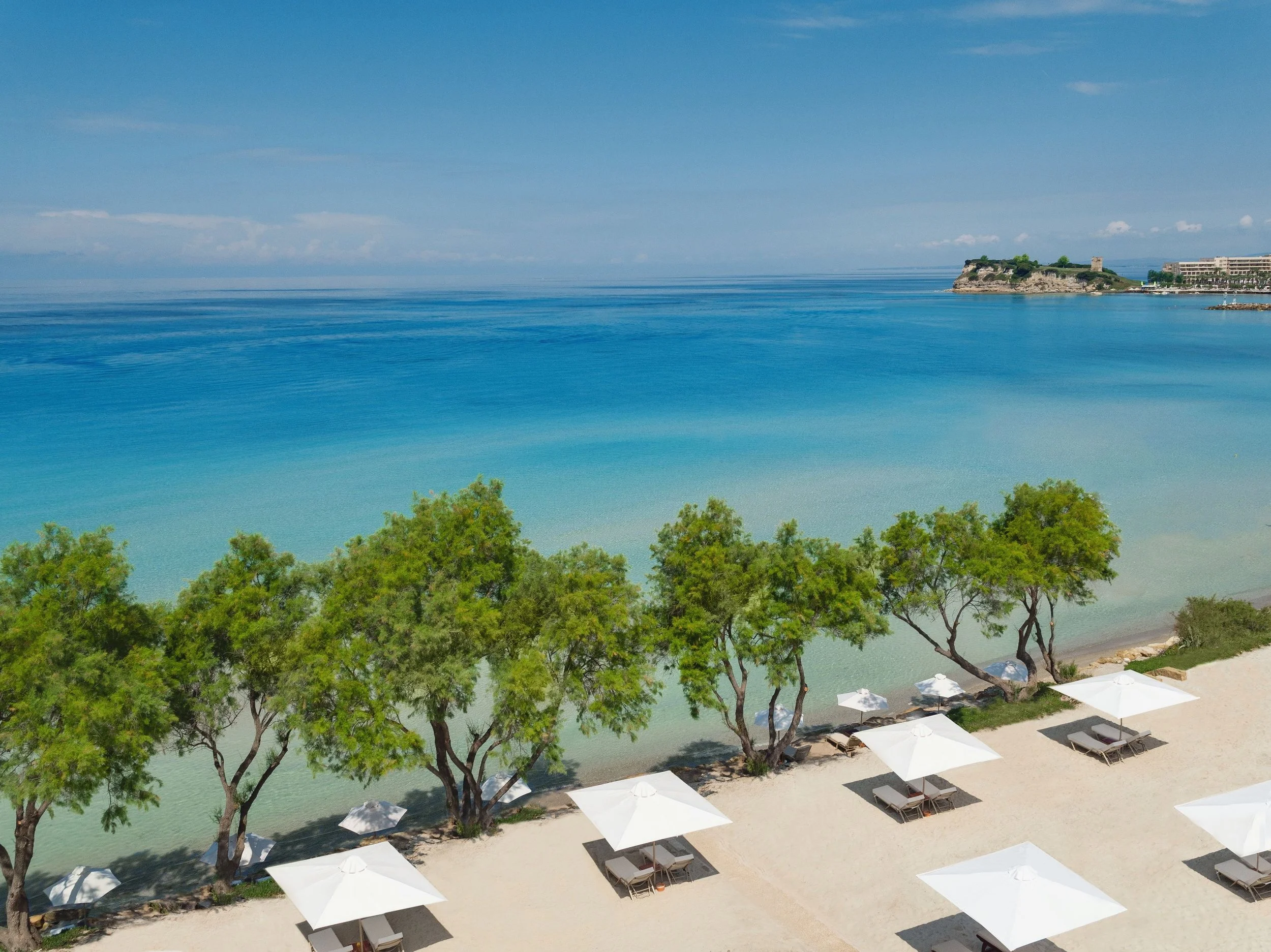 Beach with turquoise water, green trees, white umbrellas, and lounge chairs along the sandy shore, with a cliff and a building in the background under a partly cloudy sky.