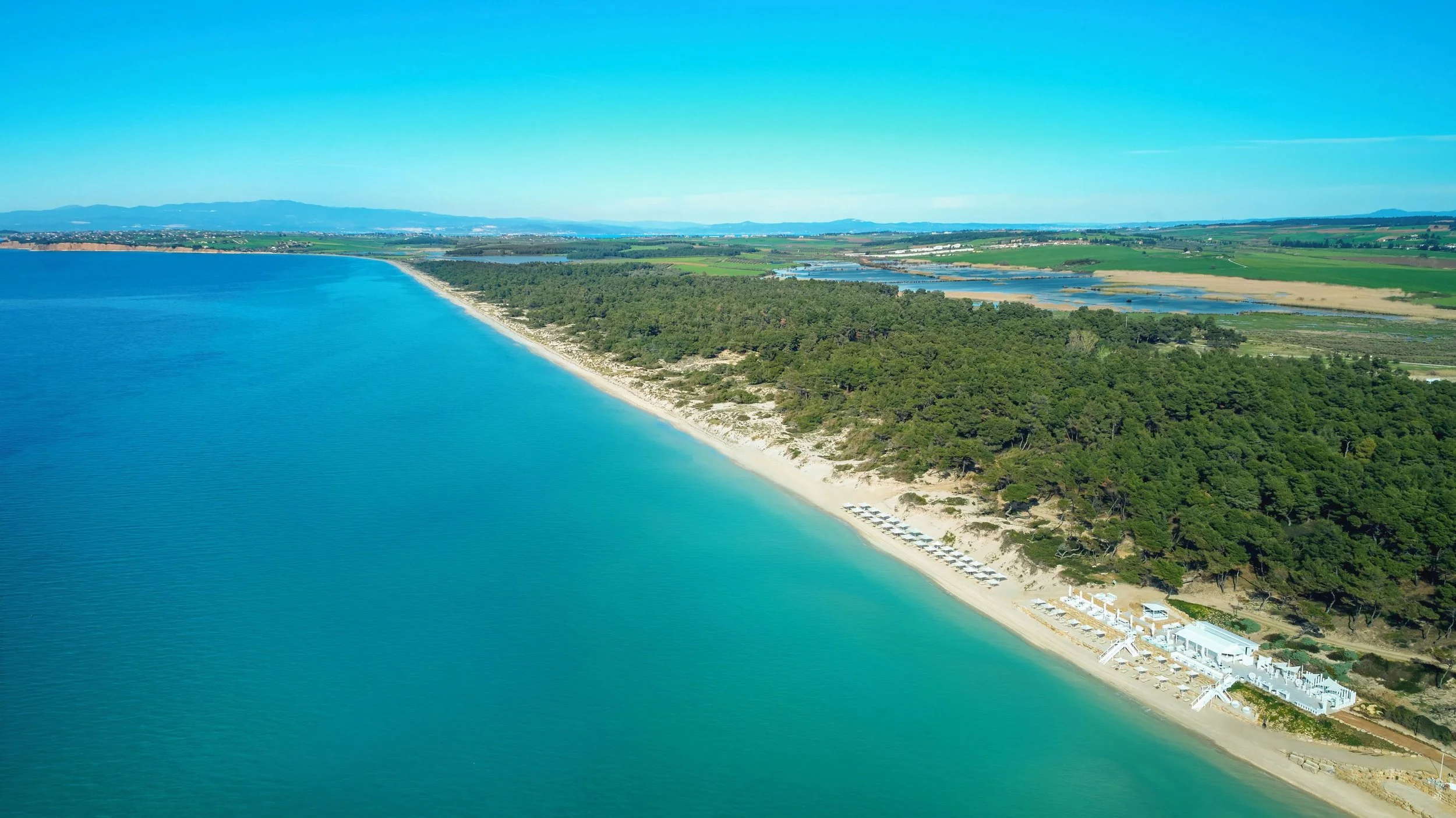 Aerial view of a coastline with a sandy beach, turquoise water, a forested area, and parked beach chairs along the shore. In the background, there are green fields and distant mountains under a clear blue sky.