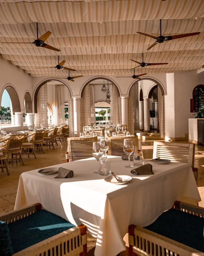 Elegant restaurant dining area with a white tablecloth-covered table set with wine glasses, napkins, and cutlery, surrounded by wooden chairs, with arched windows, drapes, and ceiling fans, bright with sunlight.