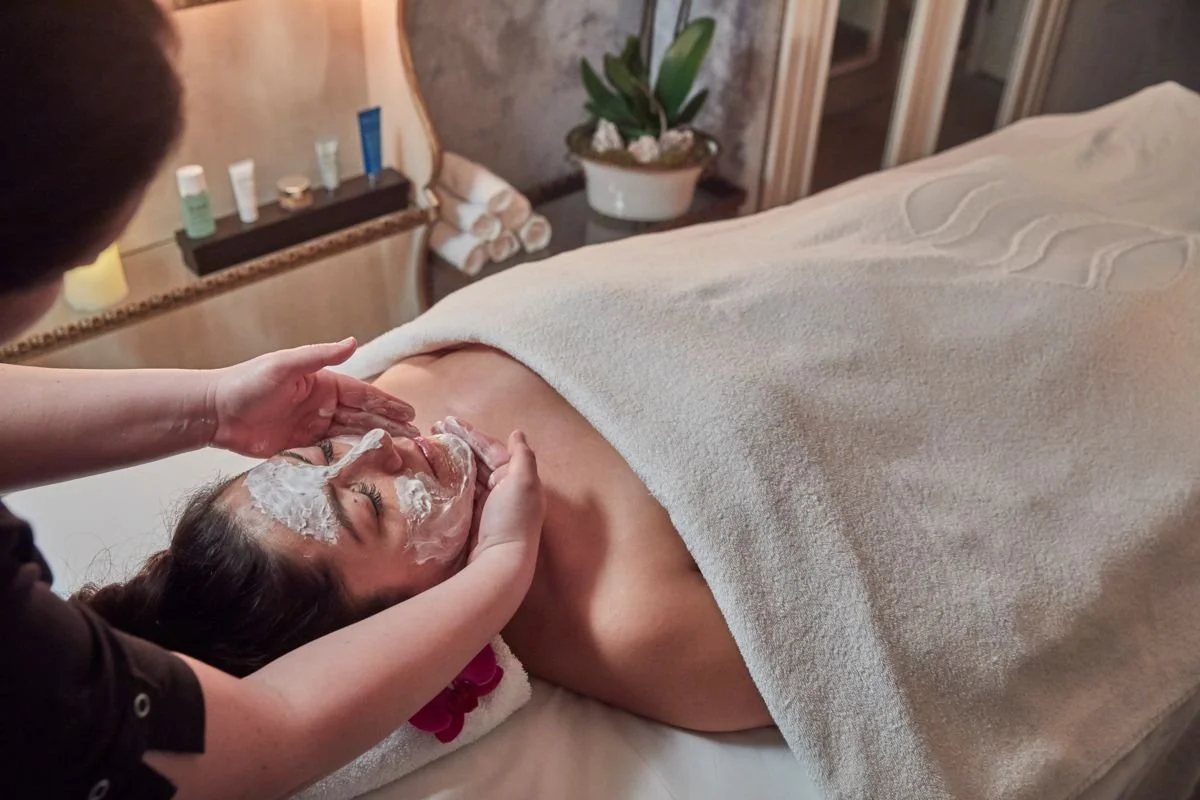 A woman receiving a facial treatment at a spa, lying on a massage table covered with a towel, while a spa professional applies a facial mask.