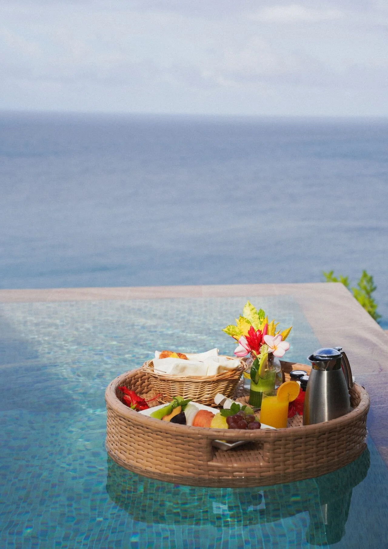 A wicker basket filled with assorted fresh fruits, a small vase with tropical flowers, a silver pitcher, and condiments, set on an outdoor table by a swimming pool with a scenic ocean view.