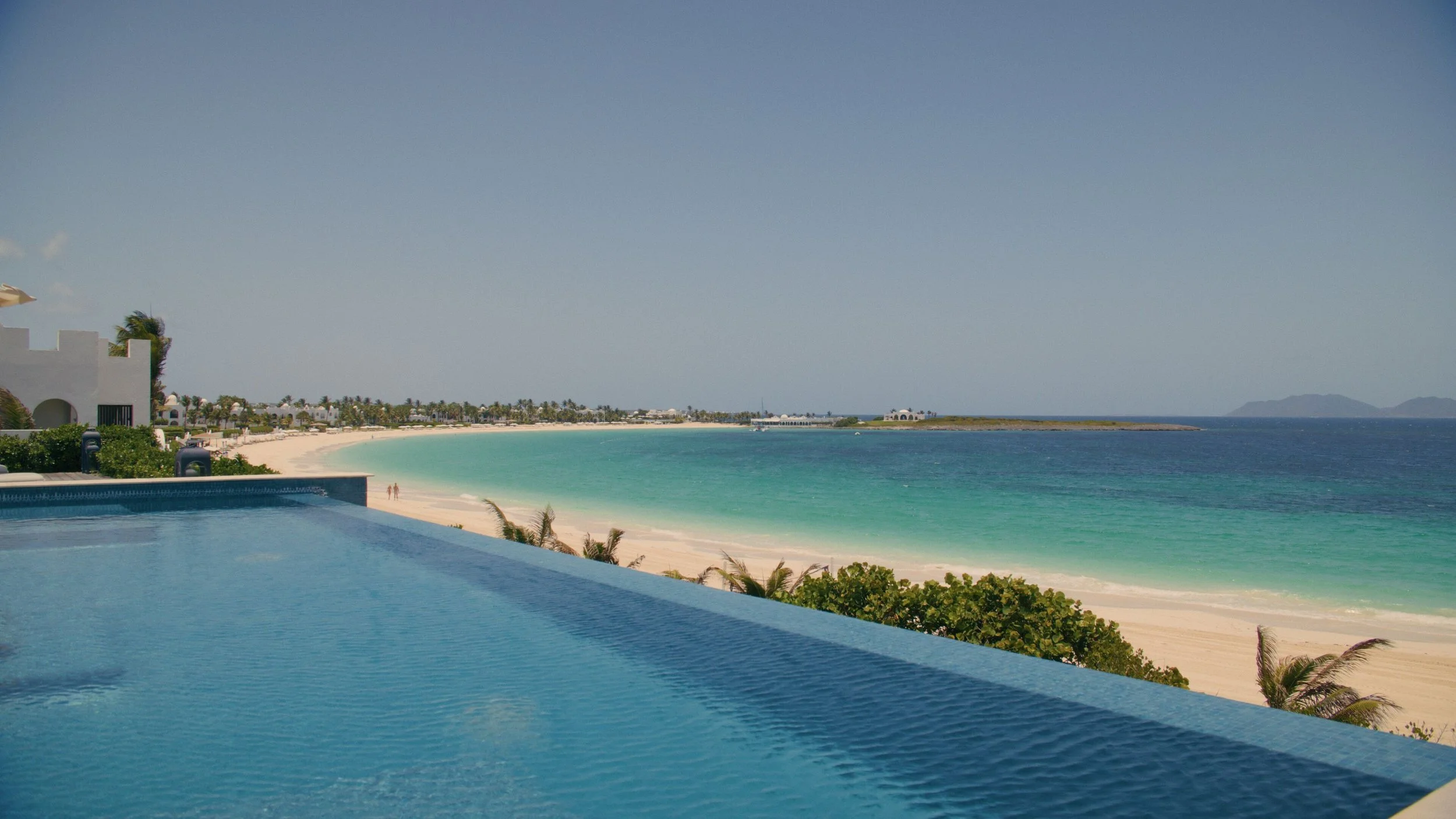 Oceanfront view with a swimming pool in the foreground, sandy beach, and turquoise water, with some buildings and trees along the shoreline under a clear blue sky.