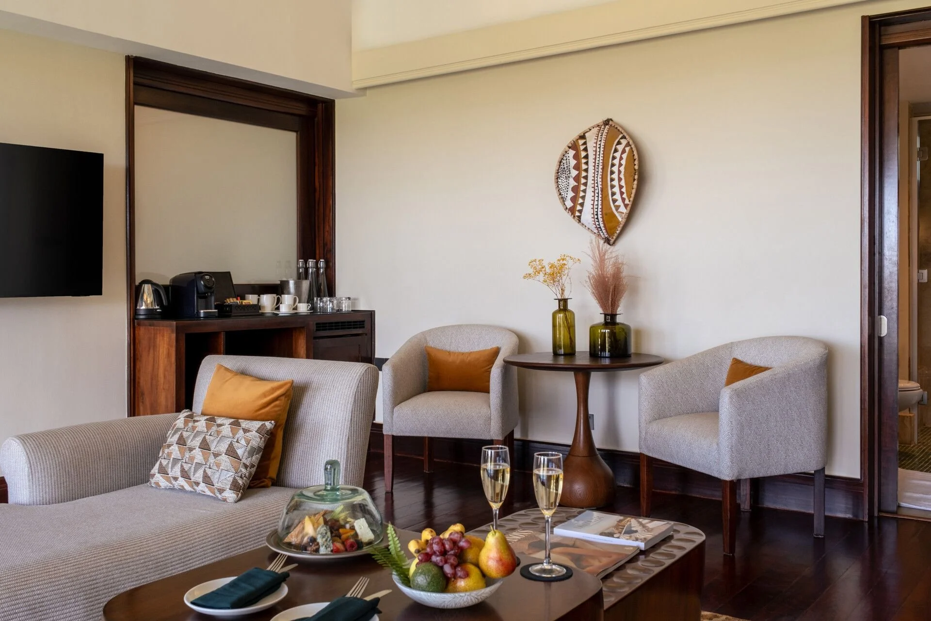 A cozy living room with two cream-colored armchairs, one with a geometric-patterned pillow, and a wooden coffee table with a fruit bowl, three glasses of champagne, and a plate with napkins. The wall is decorated with a tribal art piece and two vases with dried flowers. There is a small bar area with coffee cups, a coffee machine, and glasses.