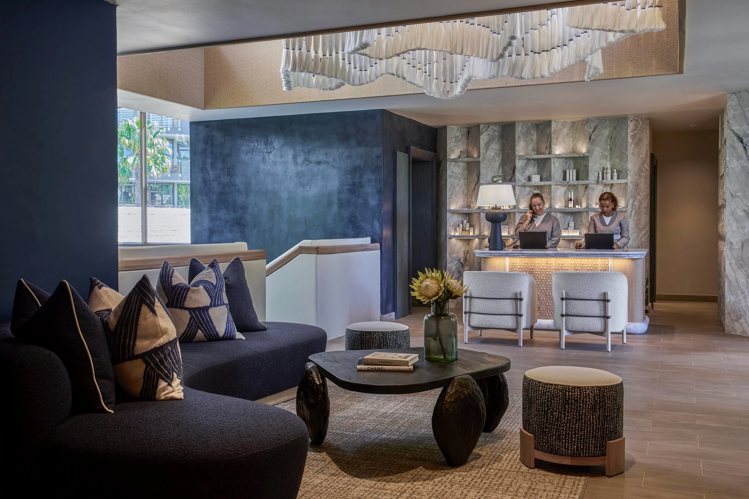 Modern hotel lobby with navy blue and beige walls, a black sofa with patterned pillows, a small tea table with books, and a marble reception desk with two smiling staff members.