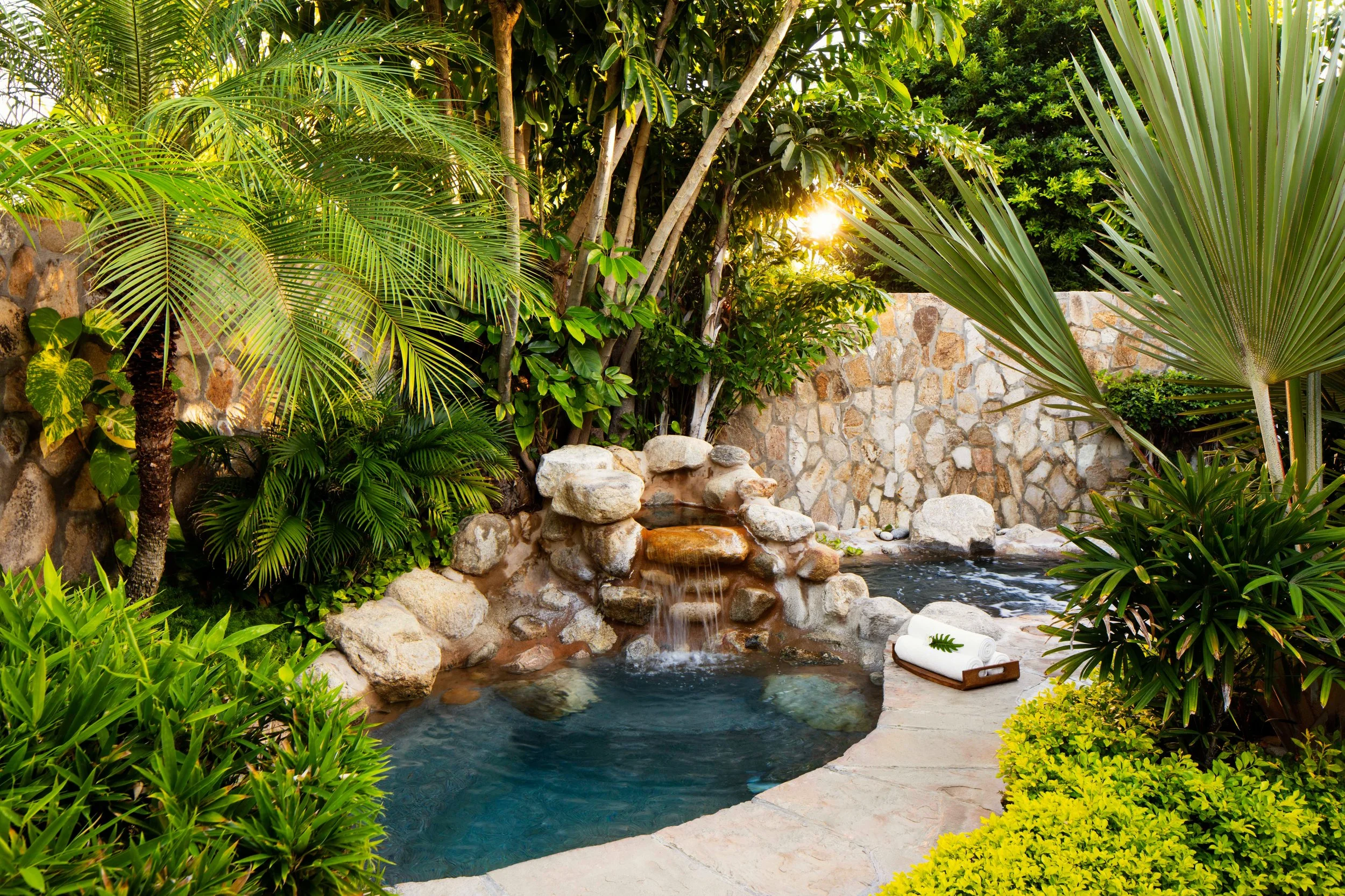 A lush tropical garden with various green plants surrounding a small pond with a waterfall feature, stone border, and some rolled towels on a tray nearby, with the sun shining through the leaves.