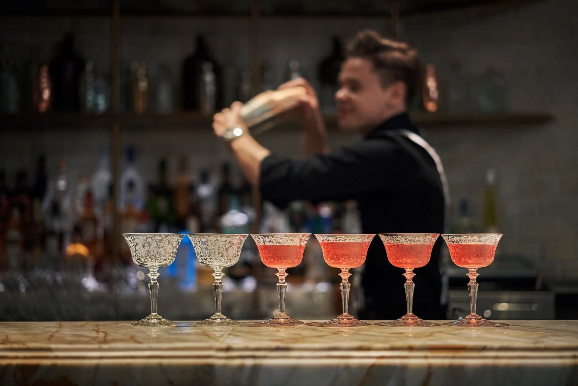 A row of six cocktail glasses with intricate floral patterns, four glasses filled with reddish-pink cocktails and two empty glasses, on a marble surface. In the background, a bartender with styled hair, wearing black clothing, is preparing drinks behind a bar with blurred bottles on shelves.