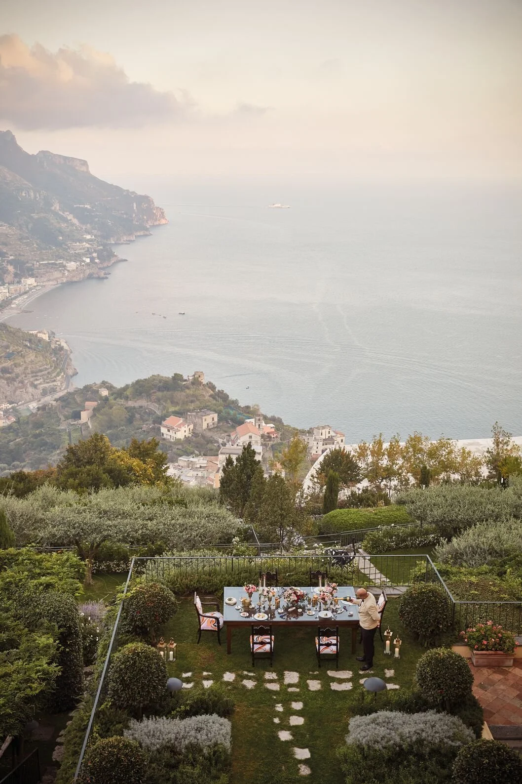 An outdoor dining setup on a lawn with a man arranging the table, overlooking a coastal village and ocean with boats, hills, and cloudy sky in the background.