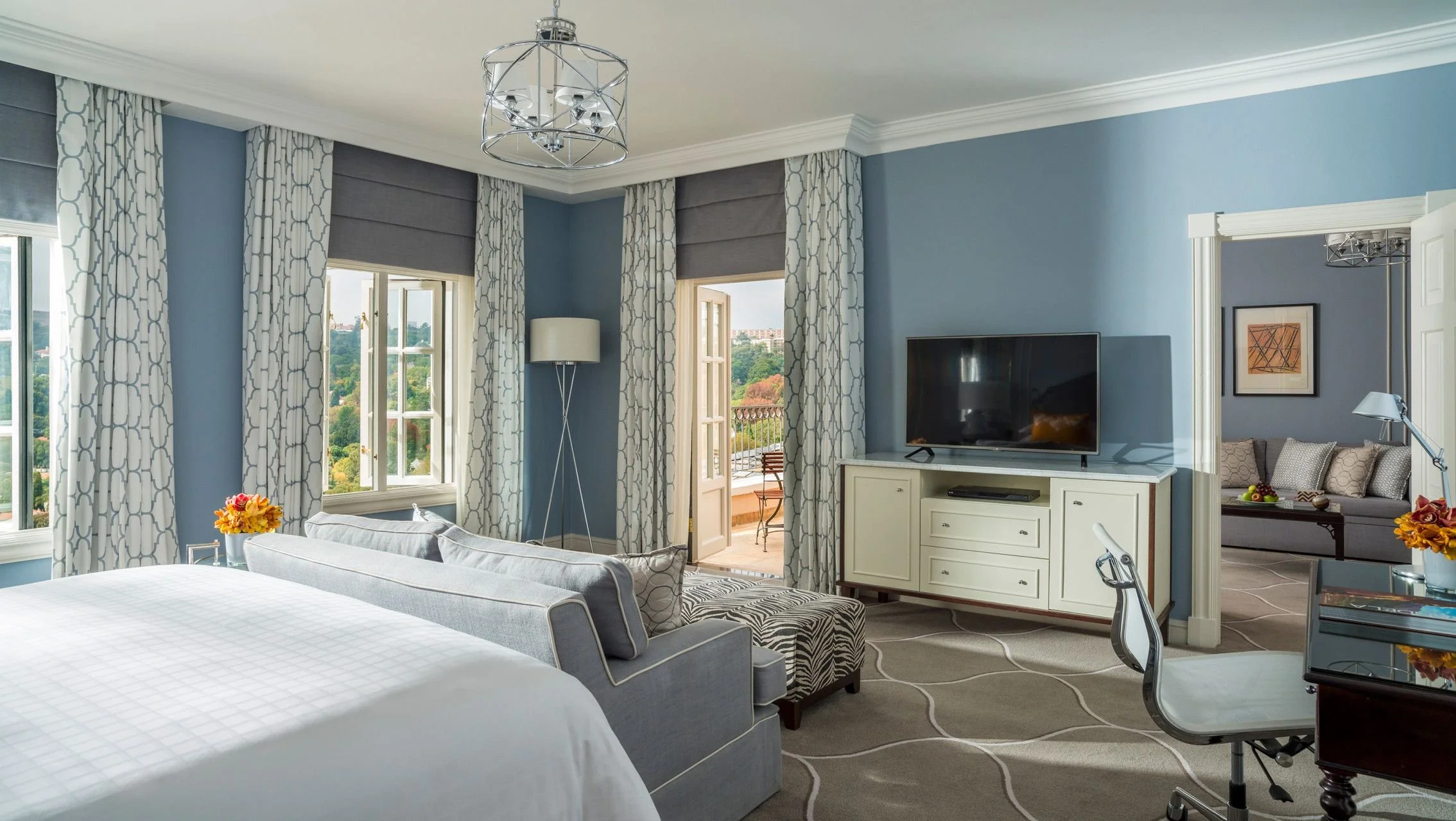 A spacious, bright hotel room with large windows, blue walls, and white decor. The room features a bed with white linens, a sitting area with a sofa, and a dresser with a flat-screen TV. French doors lead to a balcony overlooking a scenic landscape.