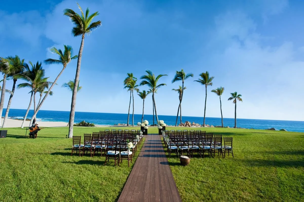 Outdoor wedding setup on a grassy lawn near the ocean, with rows of chairs, a wooden aisle, tall palm trees, and a musician with guitar