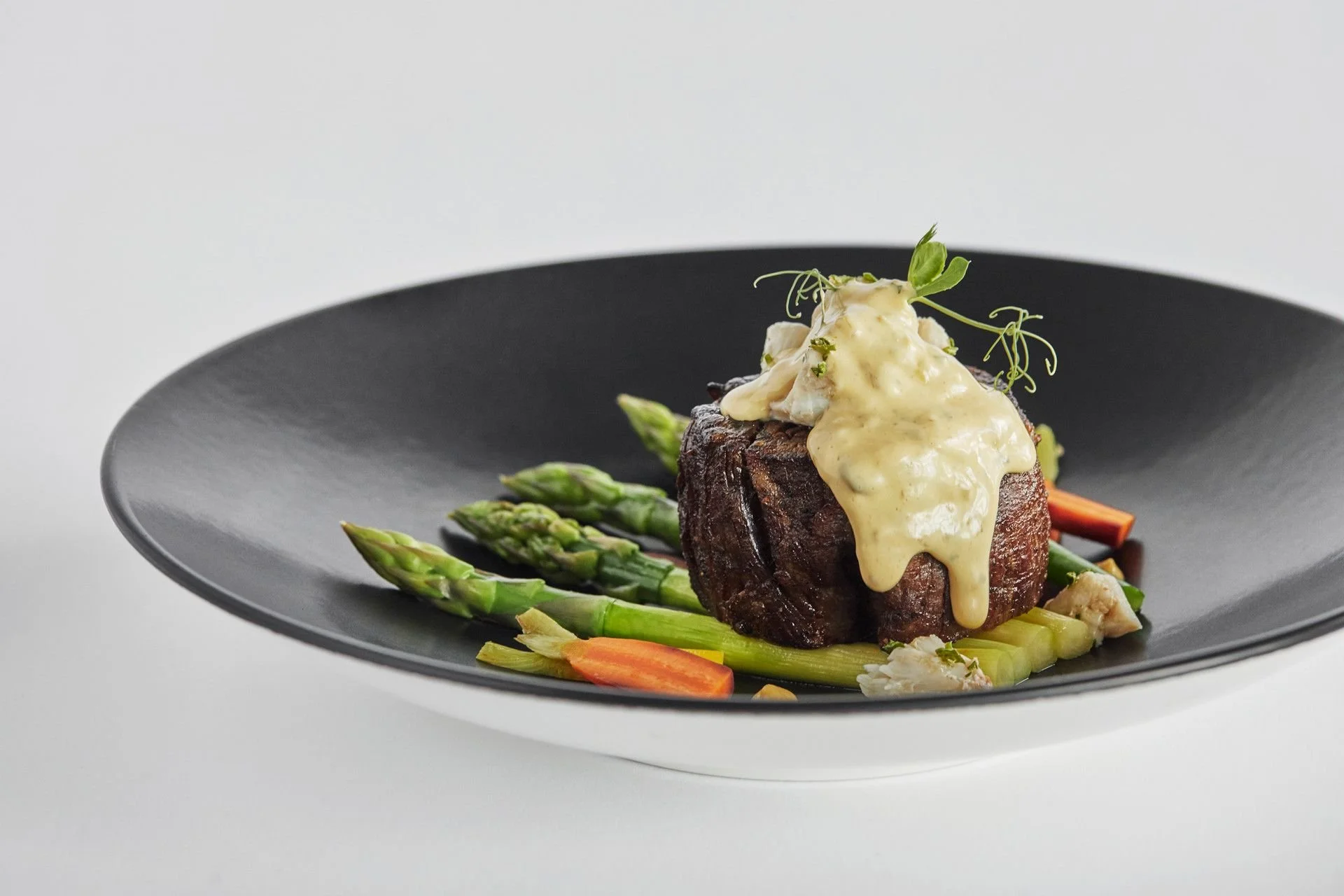 A plated gourmet dish featuring a piece of cooked steak topped with a creamy sauce, served with asparagus and vegetable garnishes on a black plate.