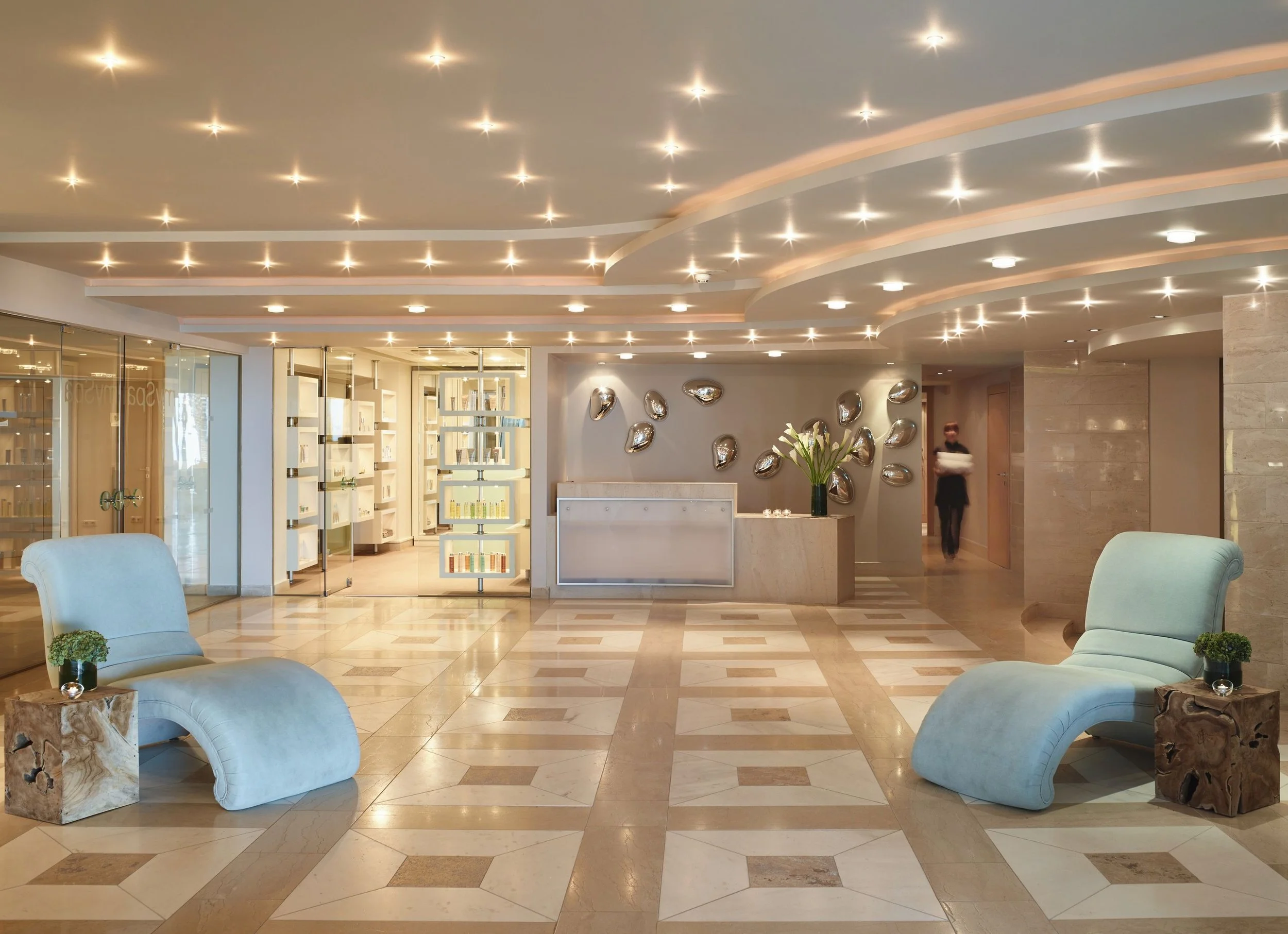 A hotel lobby with a modern design, featuring a high ceiling with numerous recessed lights, a reception desk on the right with a flower arrangement, metallic wall art, and seating area with plush chairs and side tables.