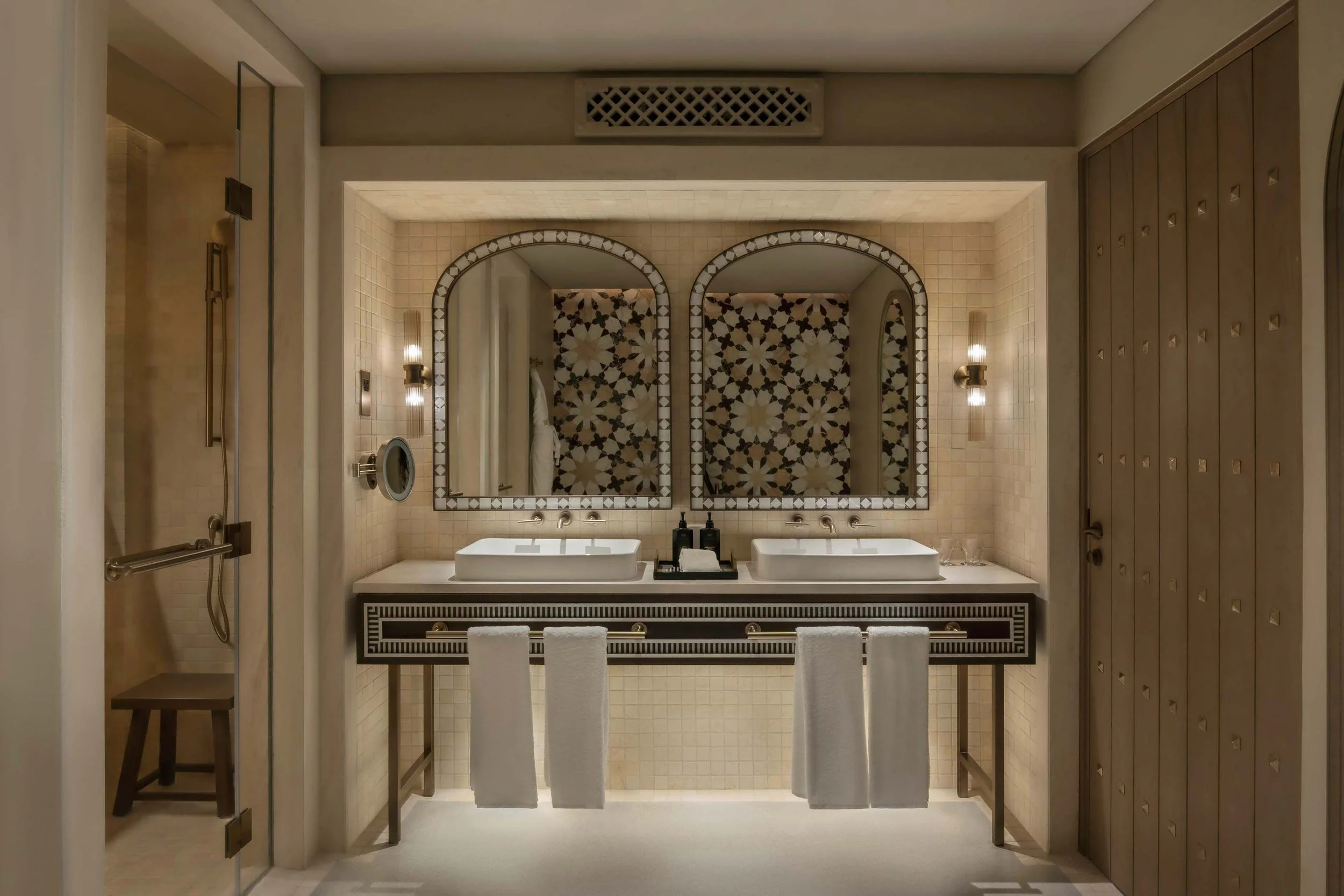 View of a luxury bathroom with two rectangular sinks, large mirrors, and a patterned tiled wall behind the sinks. The bathroom has beige tiles and decorative lighting fixtures.