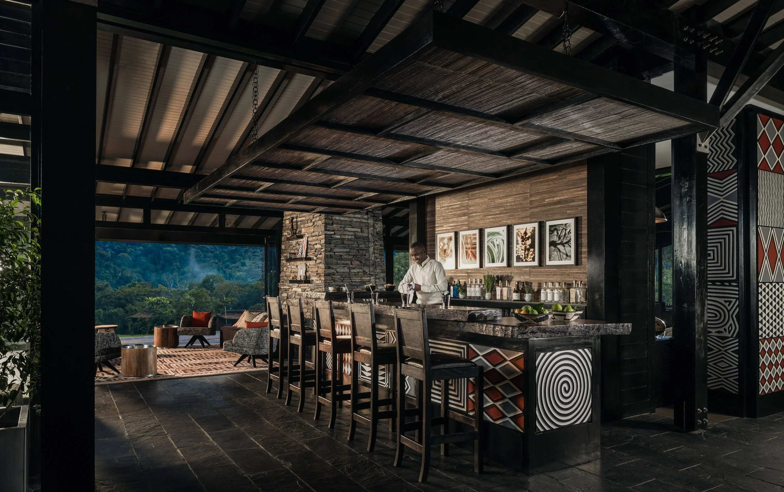 Modern living and dining area with high ceiling, black wooden beams, and large windows showcasing a scenic landscape. There is a kitchen island with a bar stool, decorated with framed art, plants, and utensils. A man in a white shirt is preparing food. Indoor seating includes a rug, chairs, and a sofa with orange pillows.