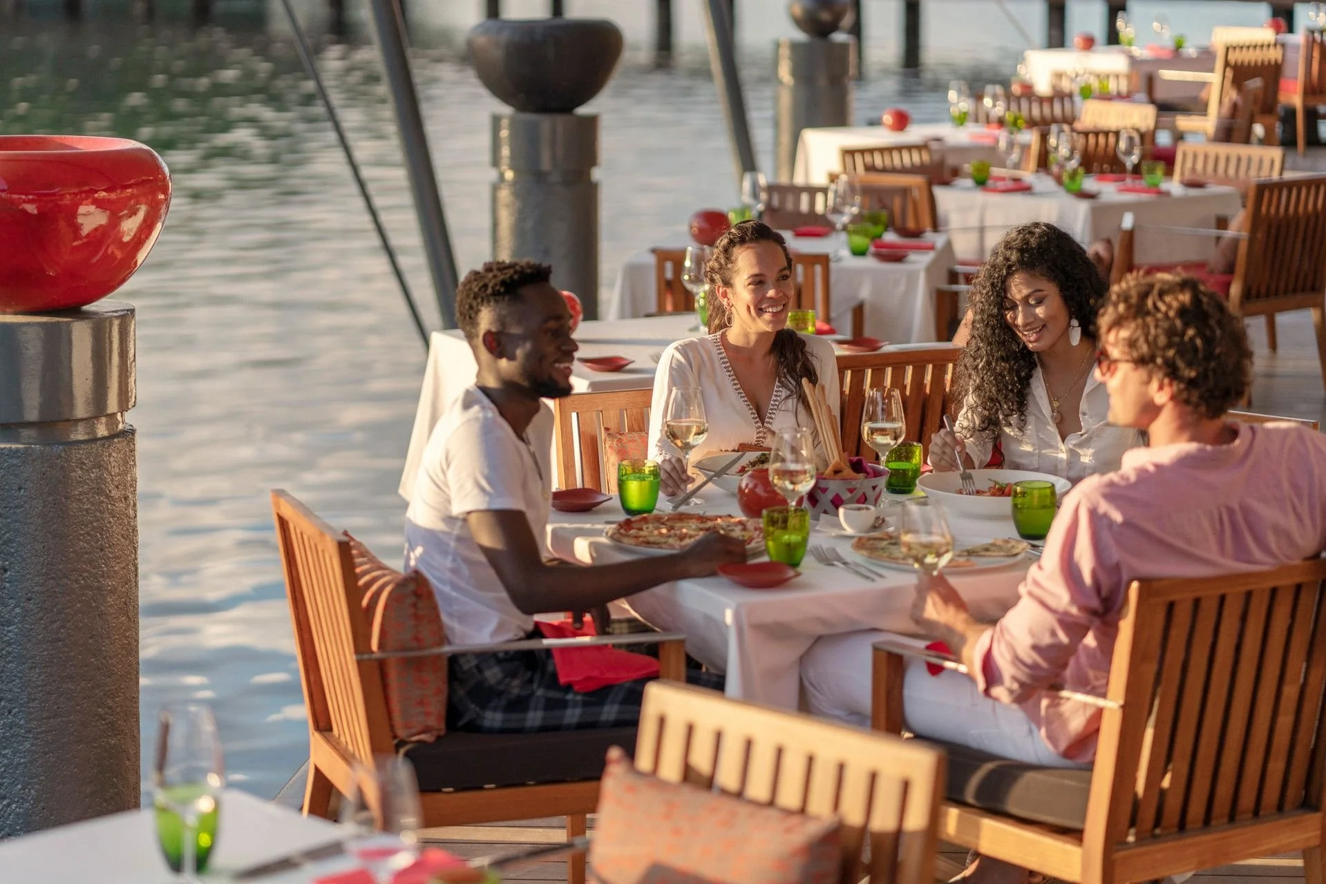 A group of four friends enjoying dinner at an outdoor restaurant by the water, with tables set with glasses, plates, and green and red tableware.