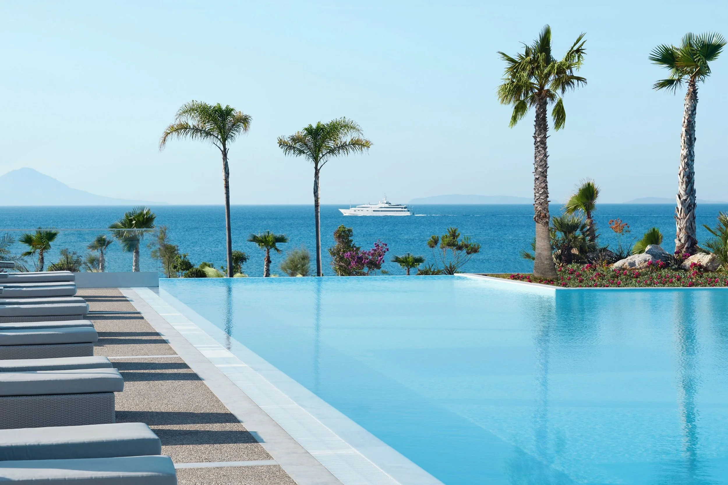 A swimming pool with lounge chairs and a view of the ocean, palm trees, and a yacht in the distance.