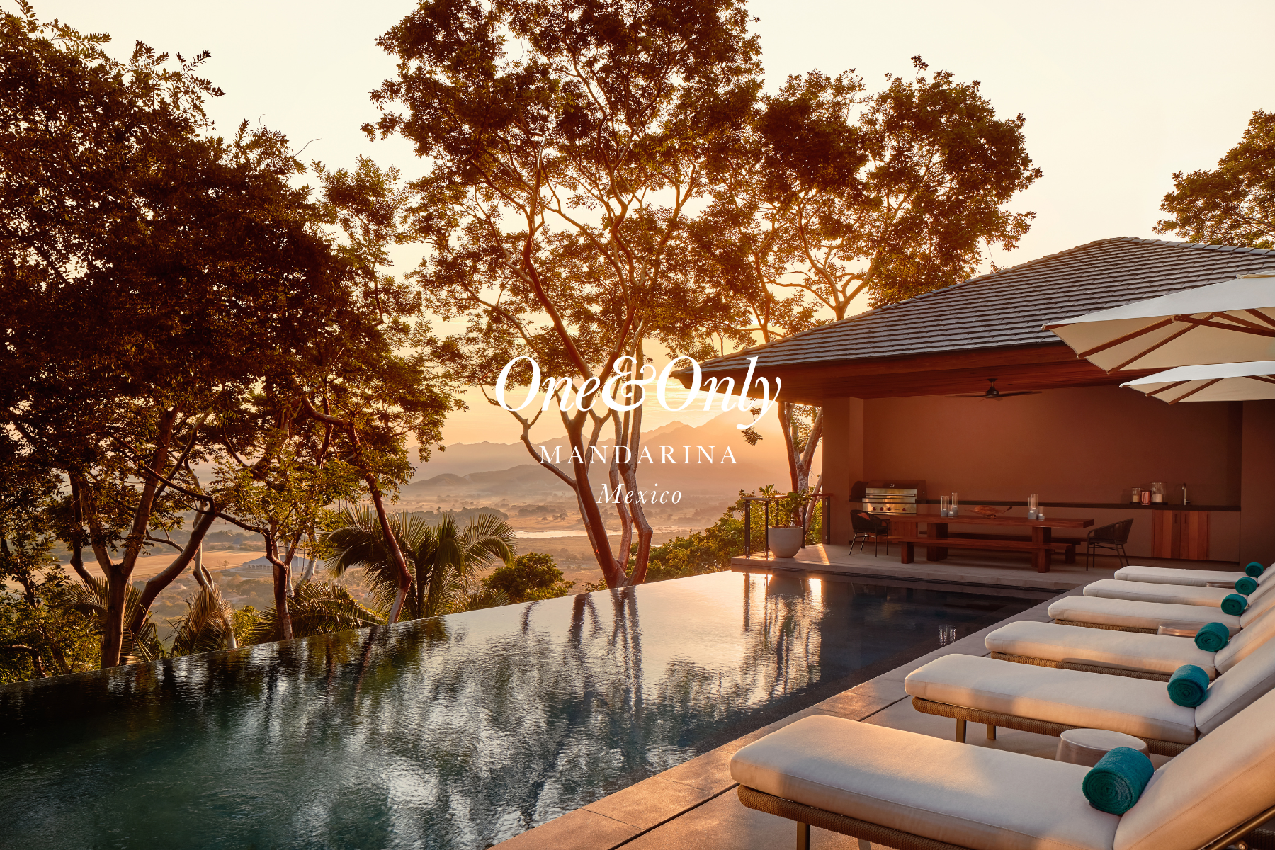 A luxurious outdoor poolside area at sunset, featuring sun loungers with towels, a modern patio with a barbecue grill, and scenic mountains in the background, in Mexico.