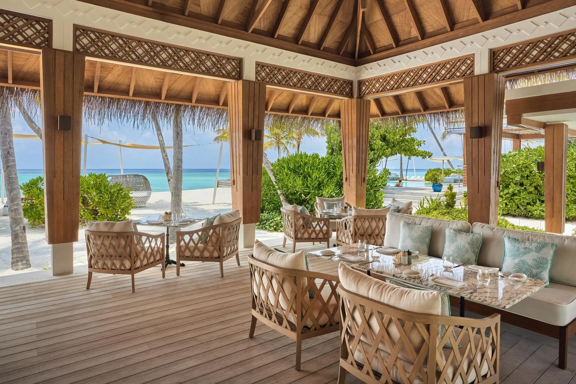 Lush outdoor dining area overlooking a beach and ocean with palm trees, under a wooden pergola, with tables, chairs, and cushions.