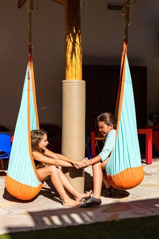 Two young girls inside hanging fabric swings, holding hands and smiling at each other, outdoors on a sunny day.